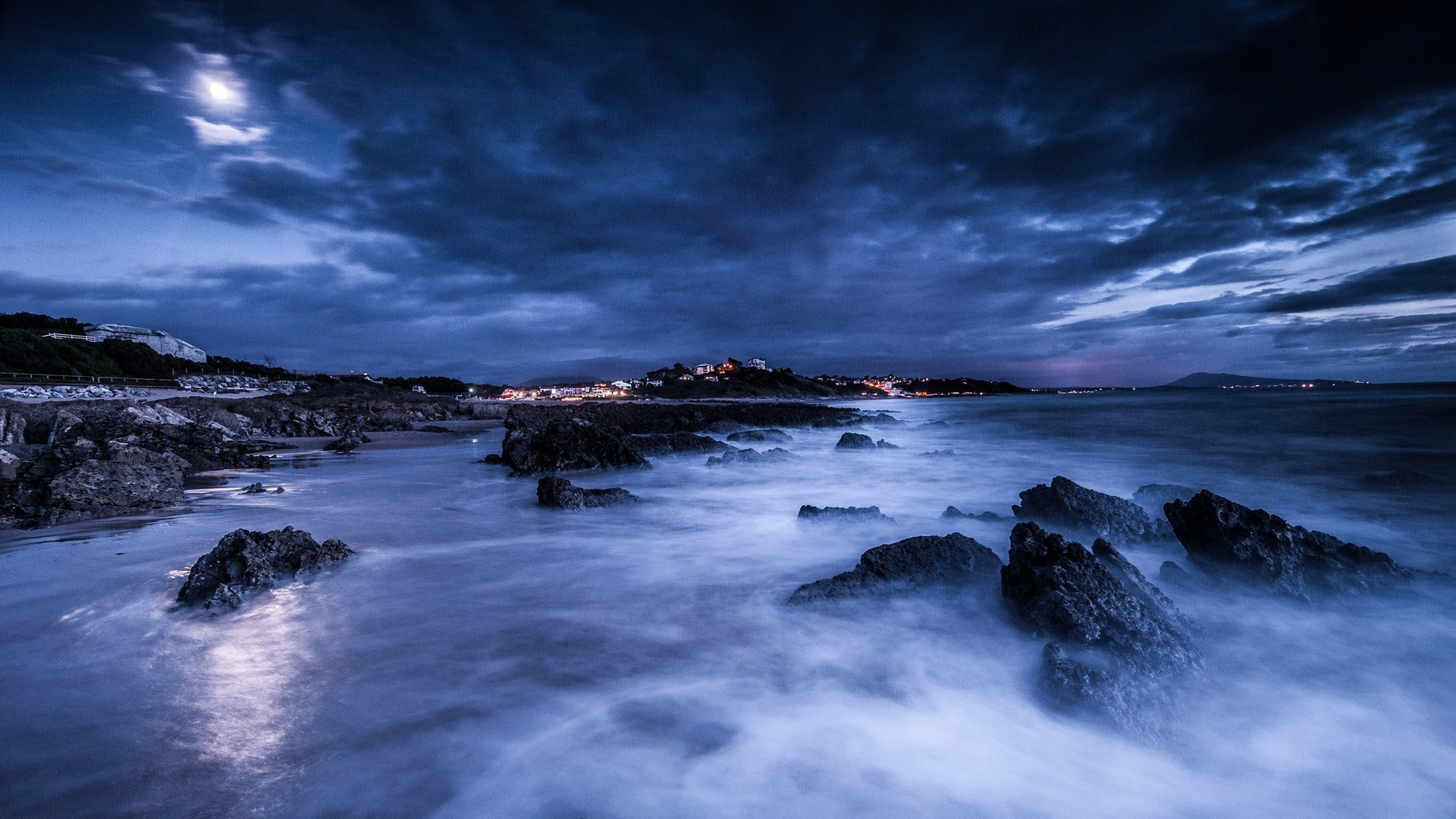 Moonlit rocky coast with waves and distant town lights HD Wallpaper