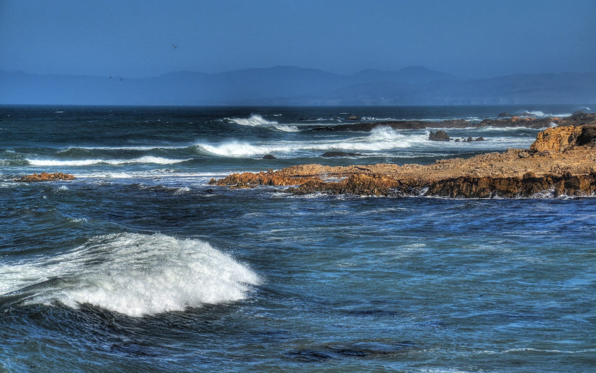 Waves crashing on rocky coastline under a blue sky HD Wallpaper