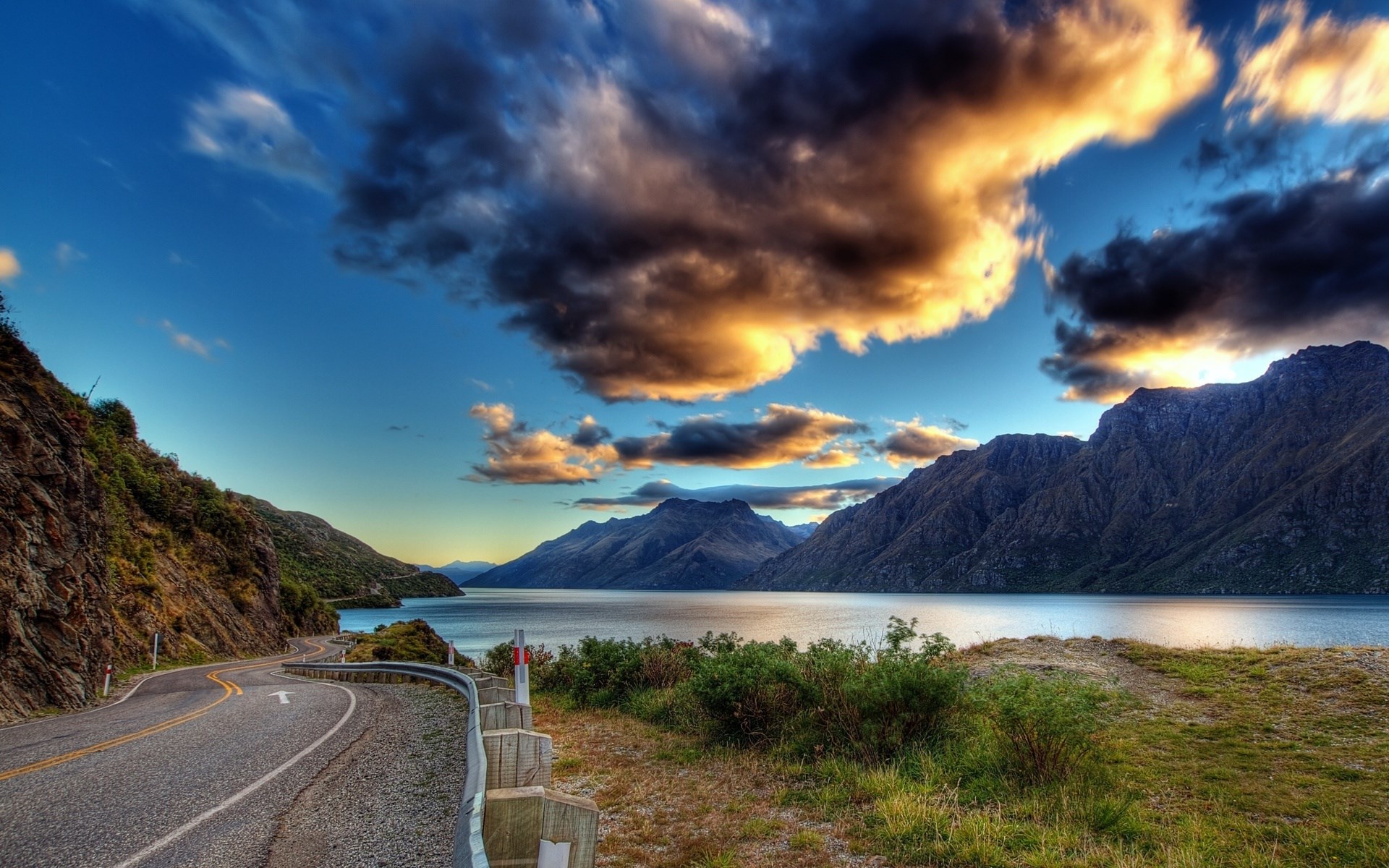 Winding road along a lake with mountains and dramatic clouds HD Wallpaper