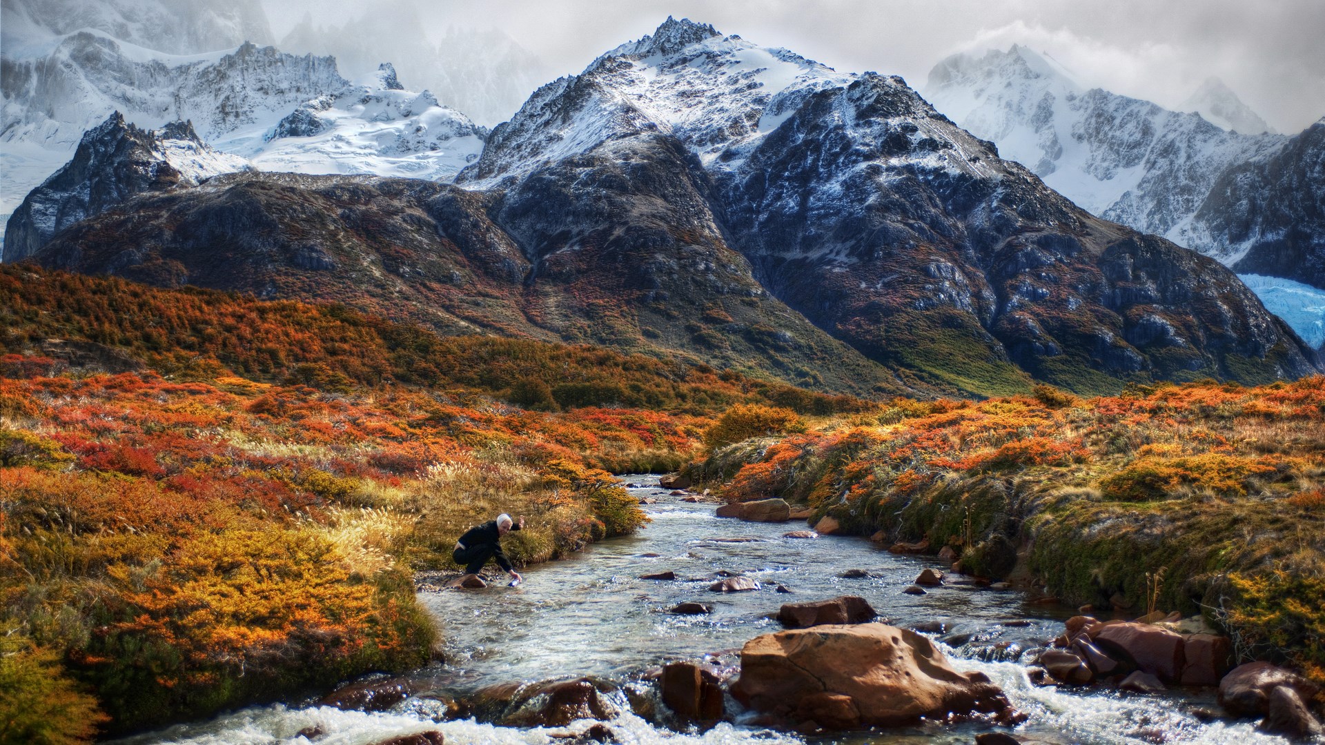 Person by stream with autumn foliage and snowy mountains HD Wallpaper