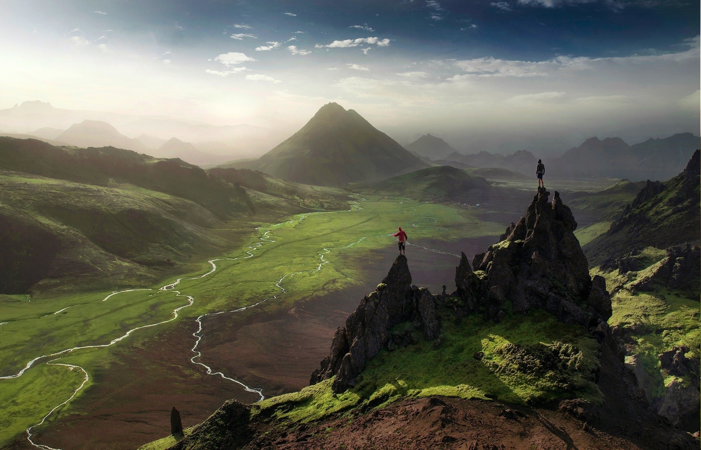 Two hikers on rocky peaks overlooking a green valley in Iceland HD Wallpaper