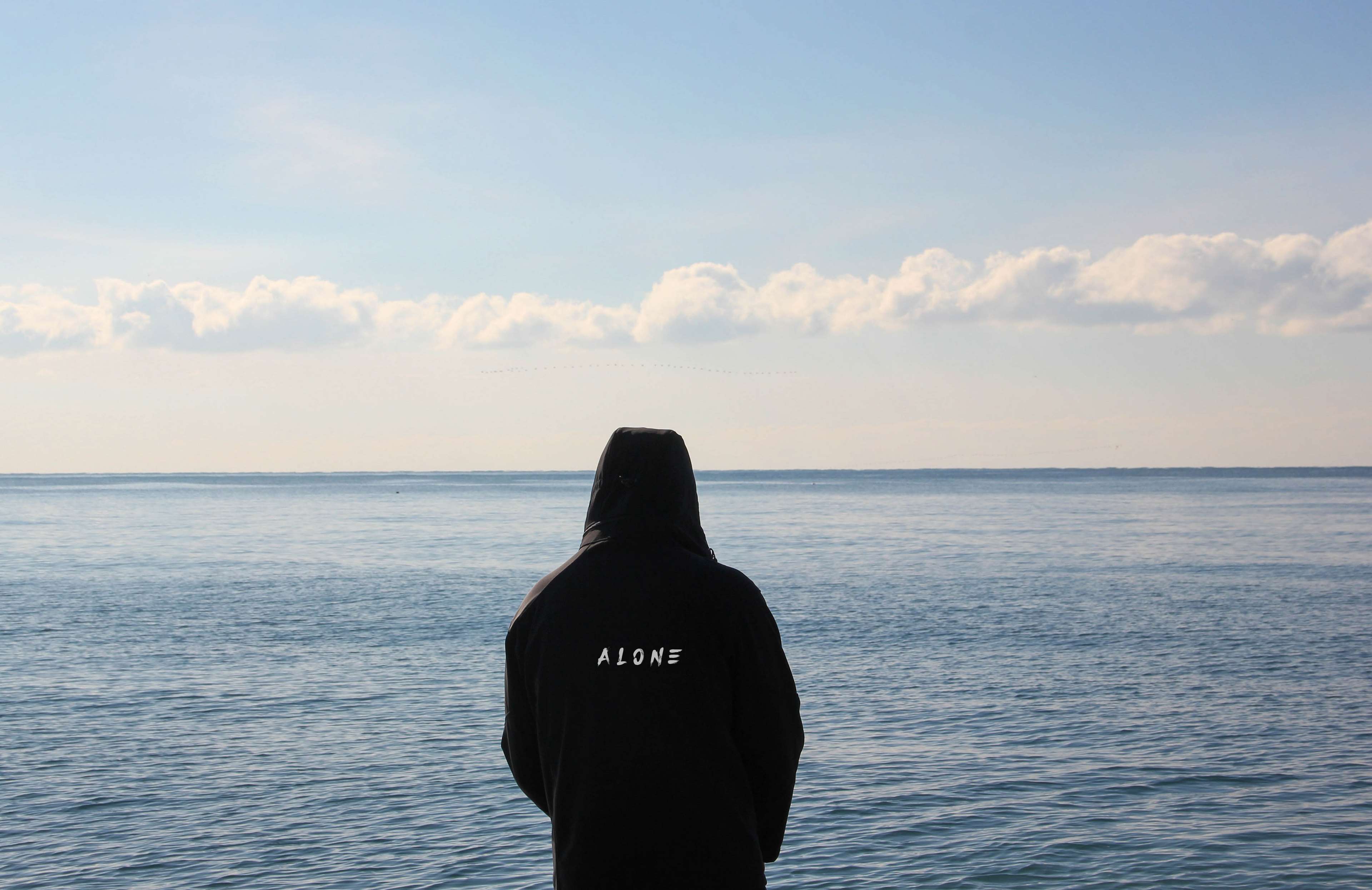 Person wearing a black hoodie with 'ALONE' text looking at the sea HD Wallpaper
