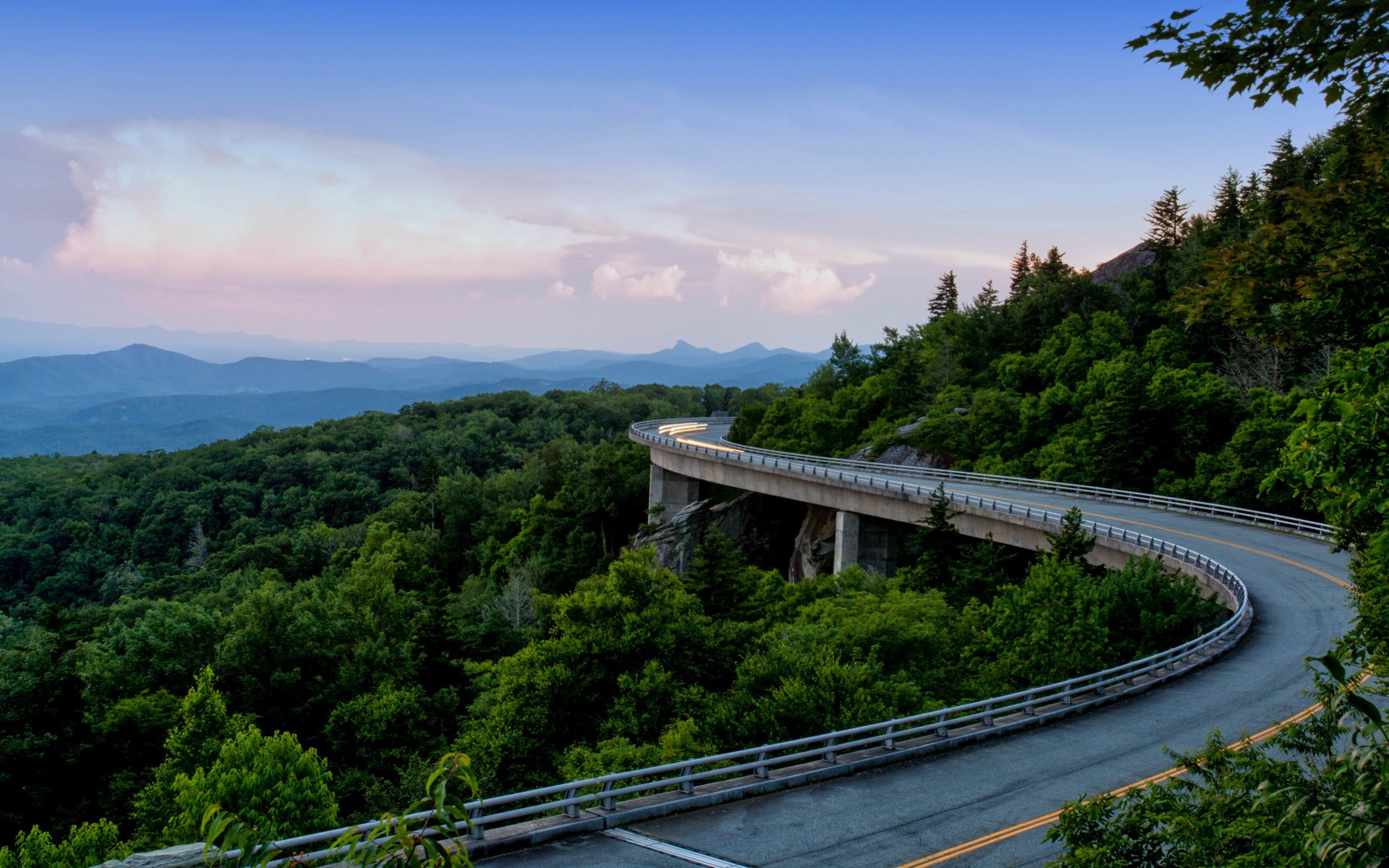 Curving bridge over a forested mountain landscape at dusk HD Wallpaper