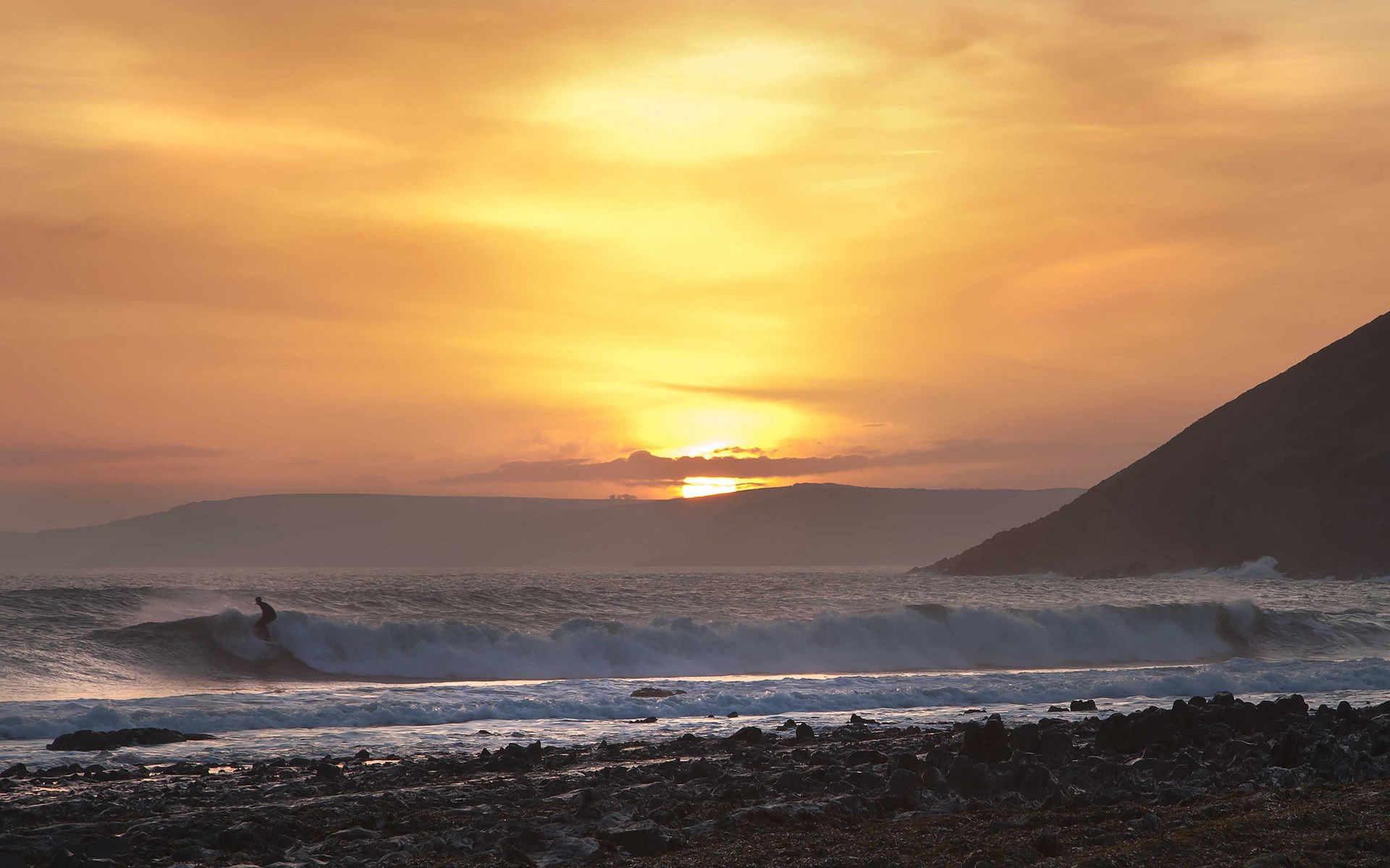Surfer riding a wave at sunset with a mountain backdrop HD Wallpaper