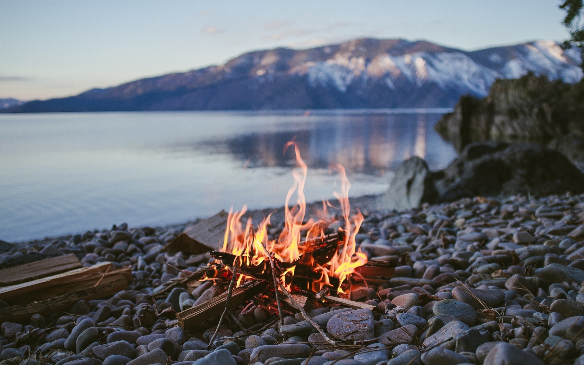Campfire on a rocky shore with a lake and mountains HD Wallpaper