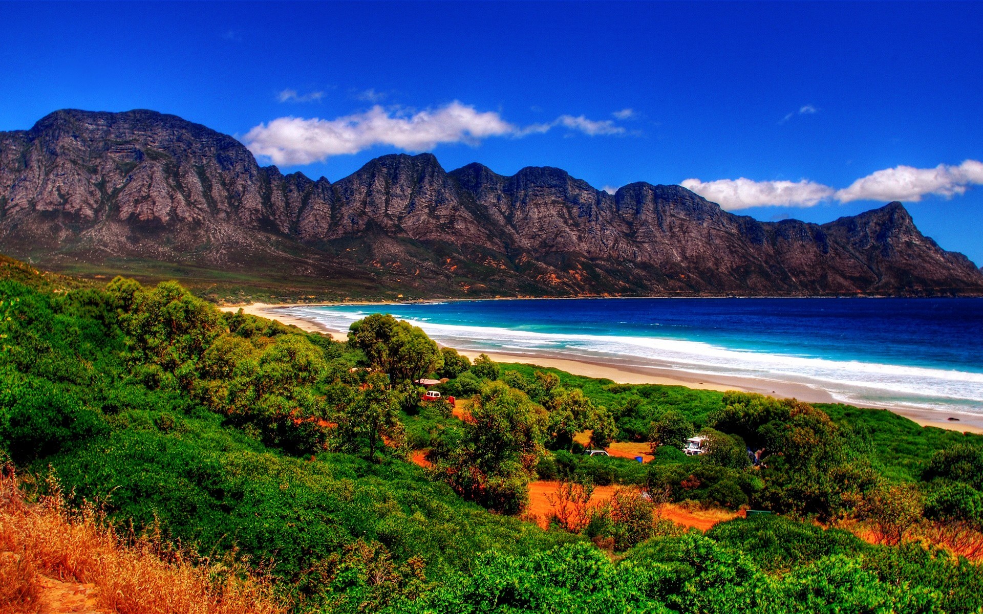 Mountain range overlooking a beach and green hills HD Wallpaper
