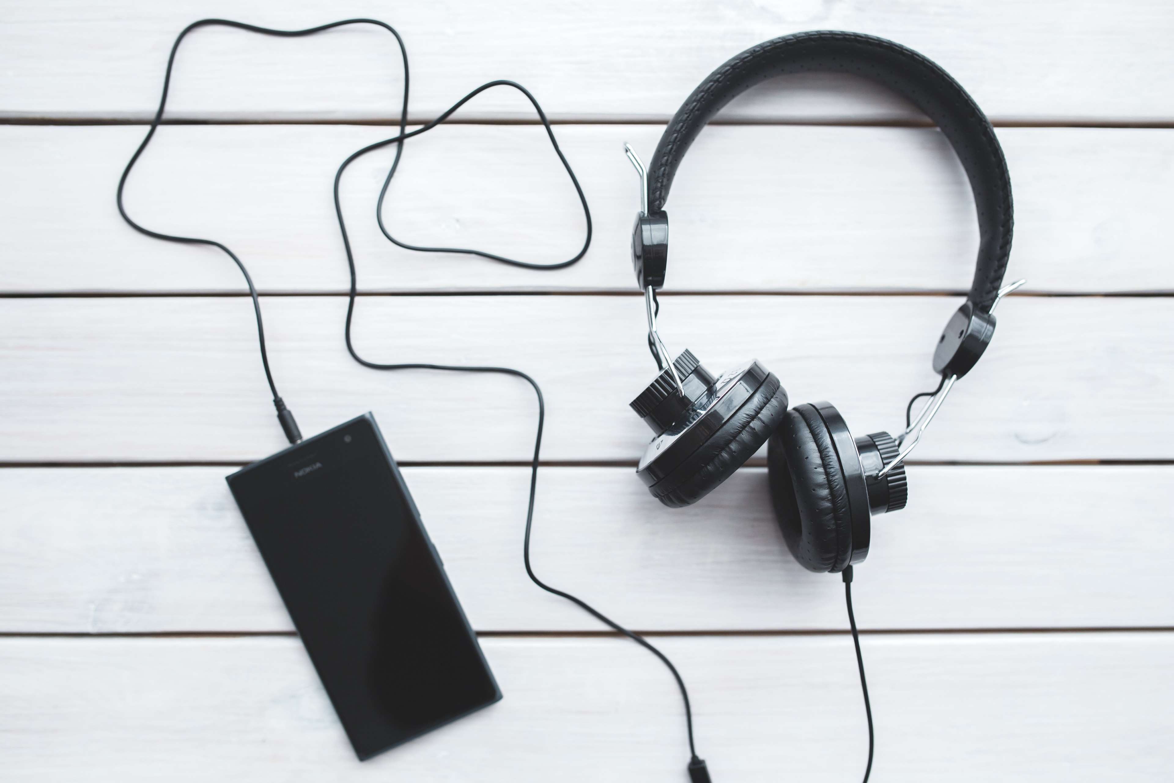 Black headphones connected to a smartphone on a white wooden surface HD Wallpaper