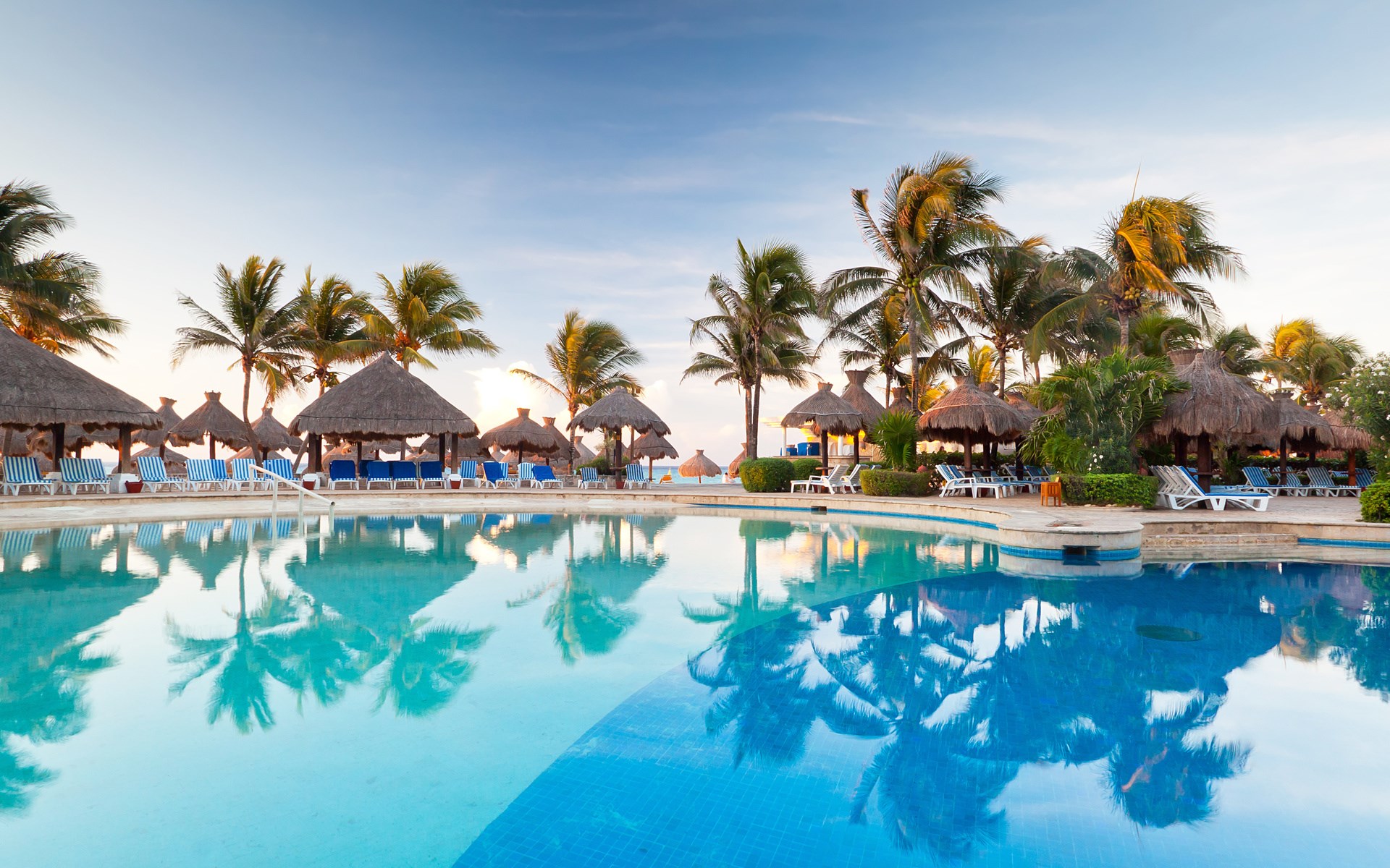 Resort swimming pool with palm trees and thatched huts HD Wallpaper