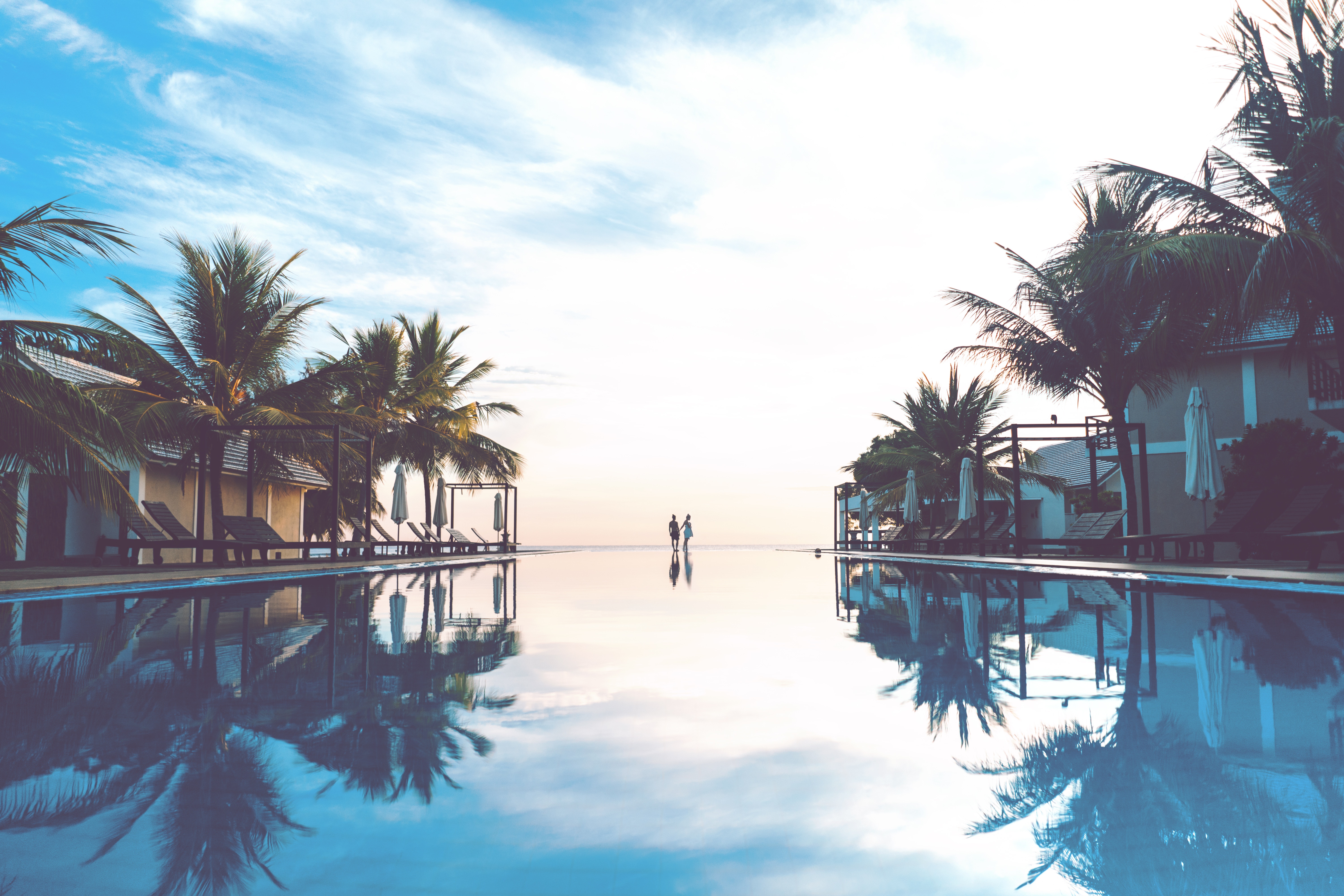 Couple walking by infinity pool with palm trees at sunset HD Wallpaper