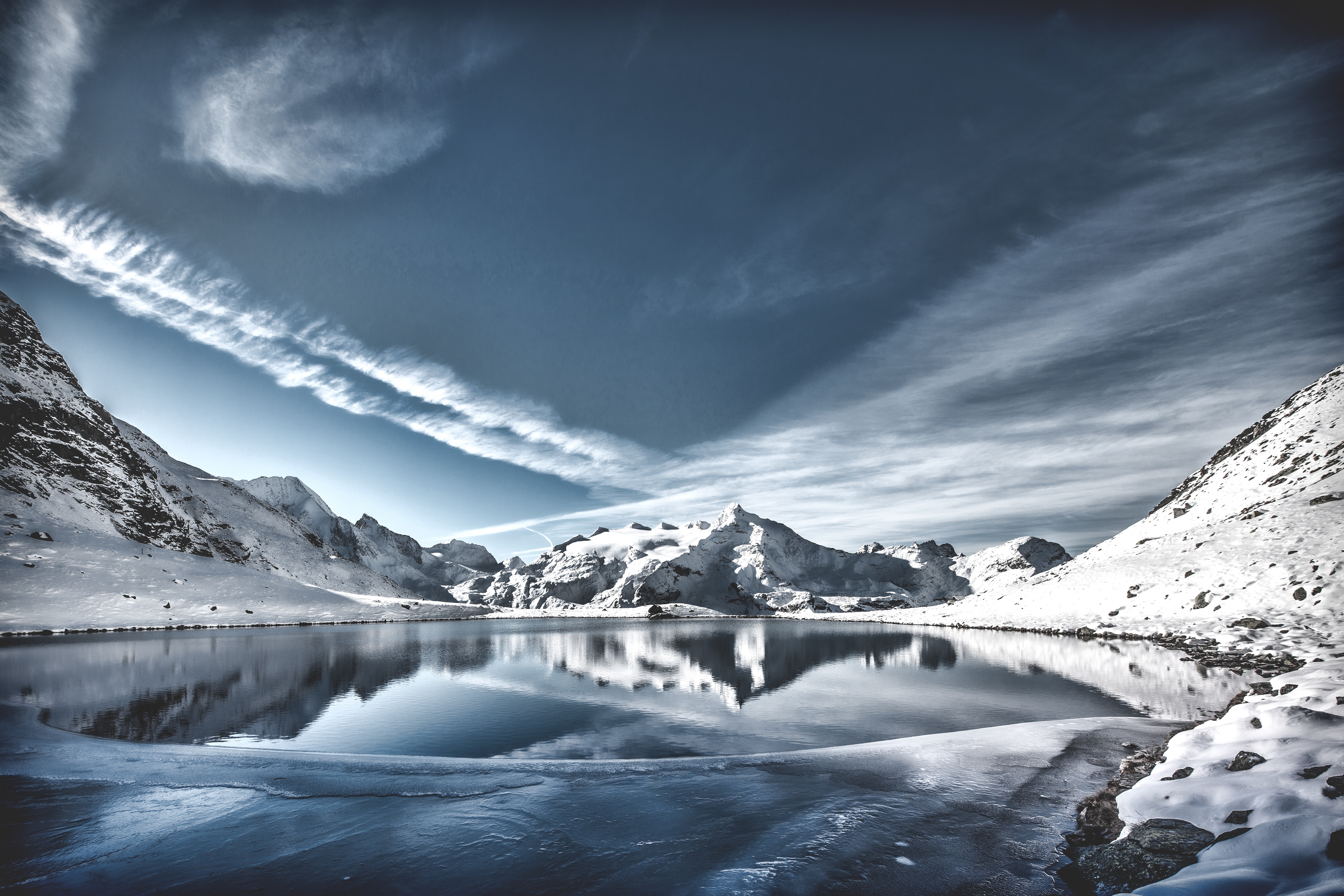 Snowy mountains reflected in a frozen lake under a cloudy sky HD Wallpaper