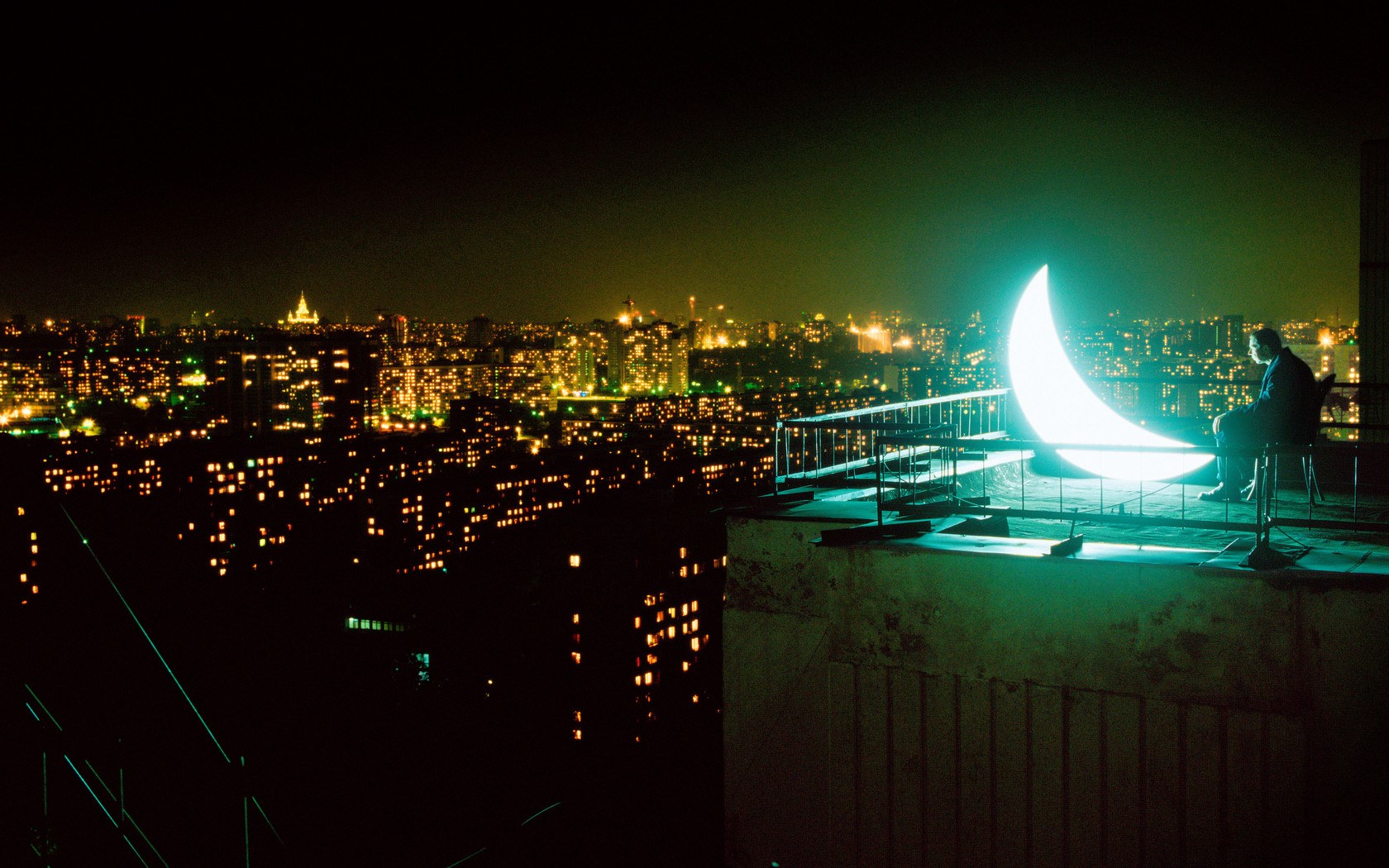Man on rooftop with glowing crescent moon overlooking city at night HD Wallpaper