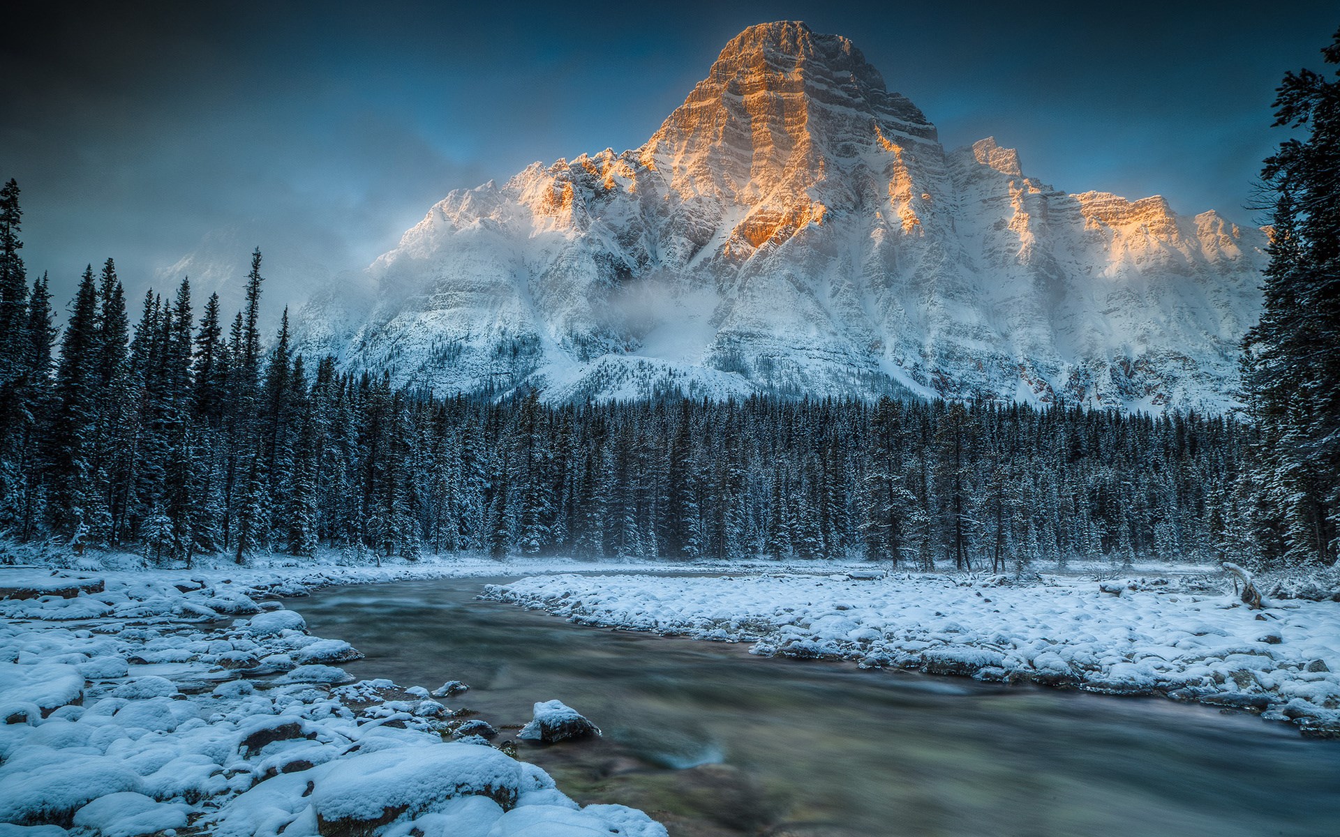 Snowy mountain peak illuminated by golden sunlight over a frozen forest and river HD Wallpaper