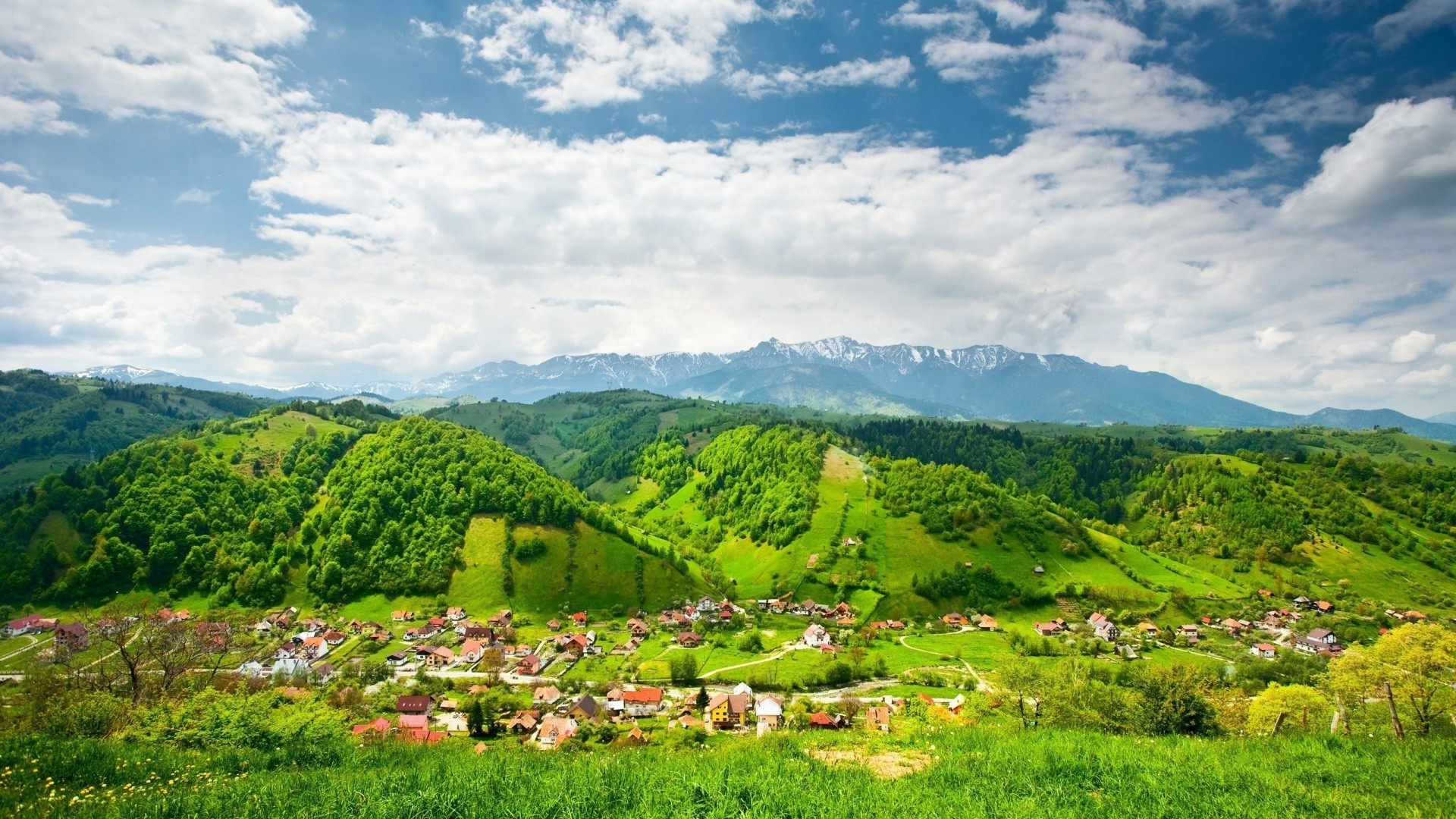 Village nestled in green hills with mountains under cloudy sky HD Wallpaper
