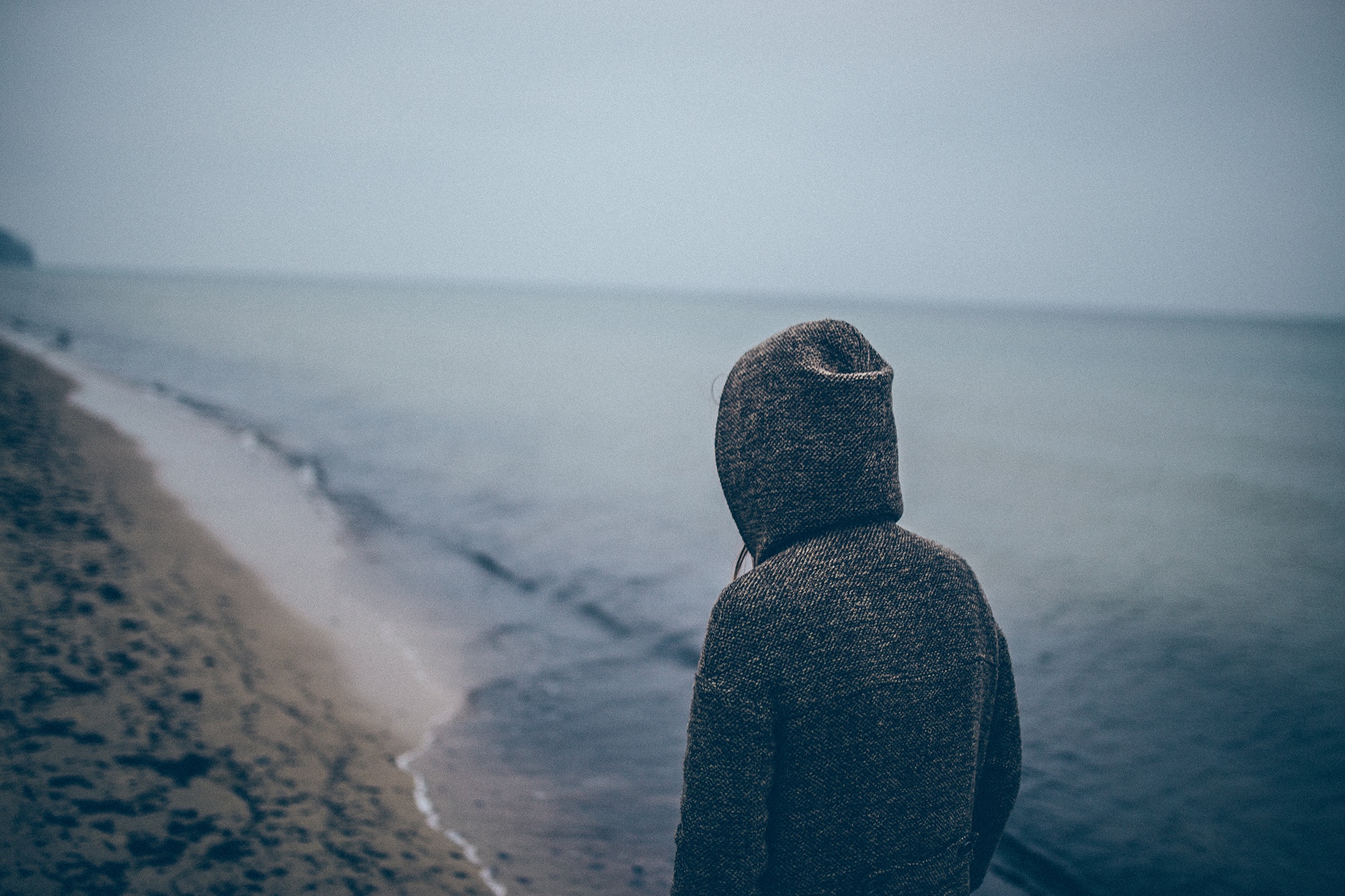 Person in hoodie looking out at the sea from a beach HD Wallpaper