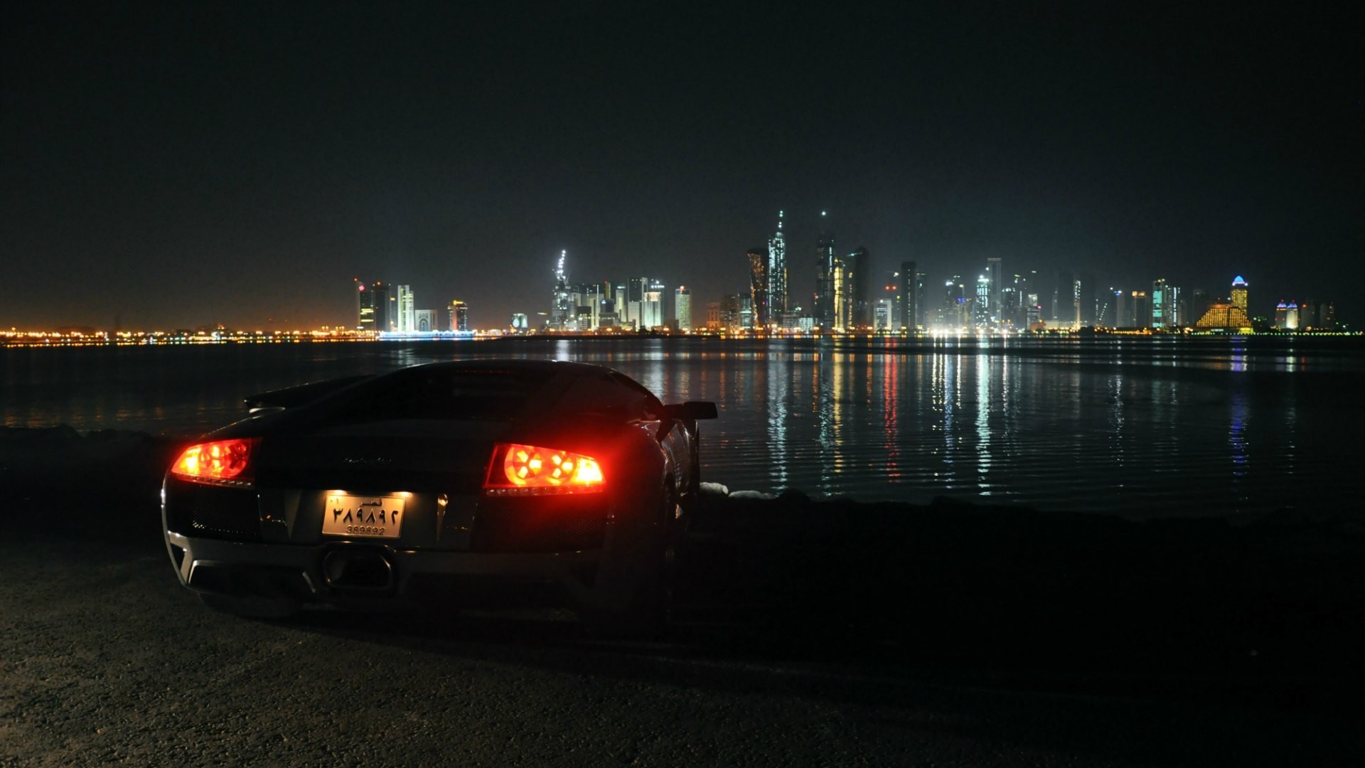 Black Lamborghini parked by the water at night with city skyline HD Wallpaper