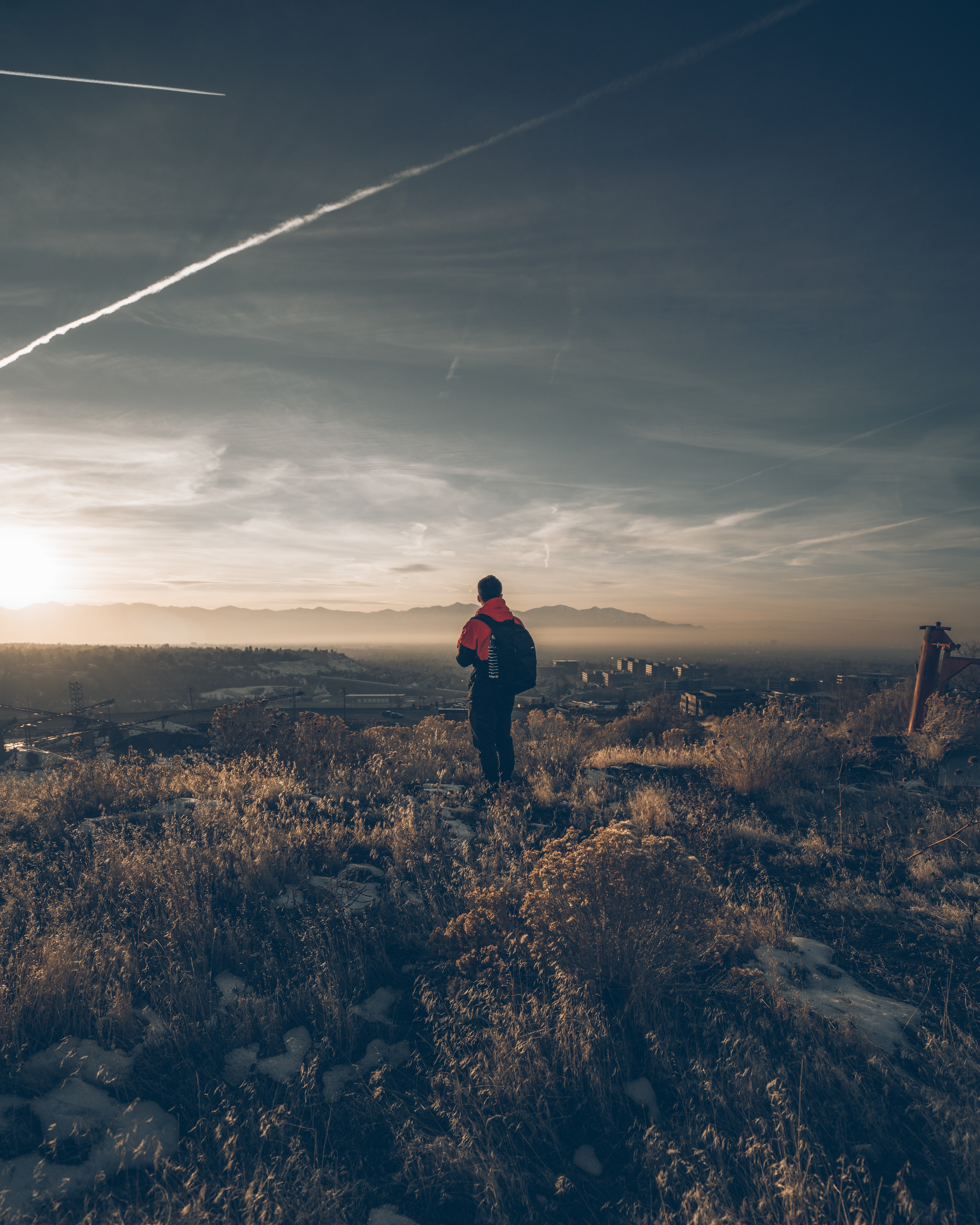 Person with backpack on grassy hill overlooking city at sunset HD Wallpaper