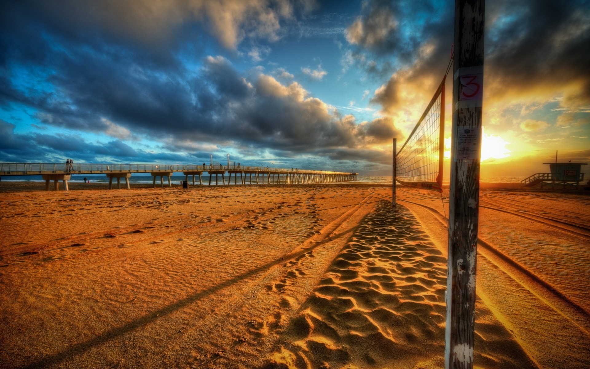 Beach volleyball net at sunset with pier and lifeguard tower HD Wallpaper