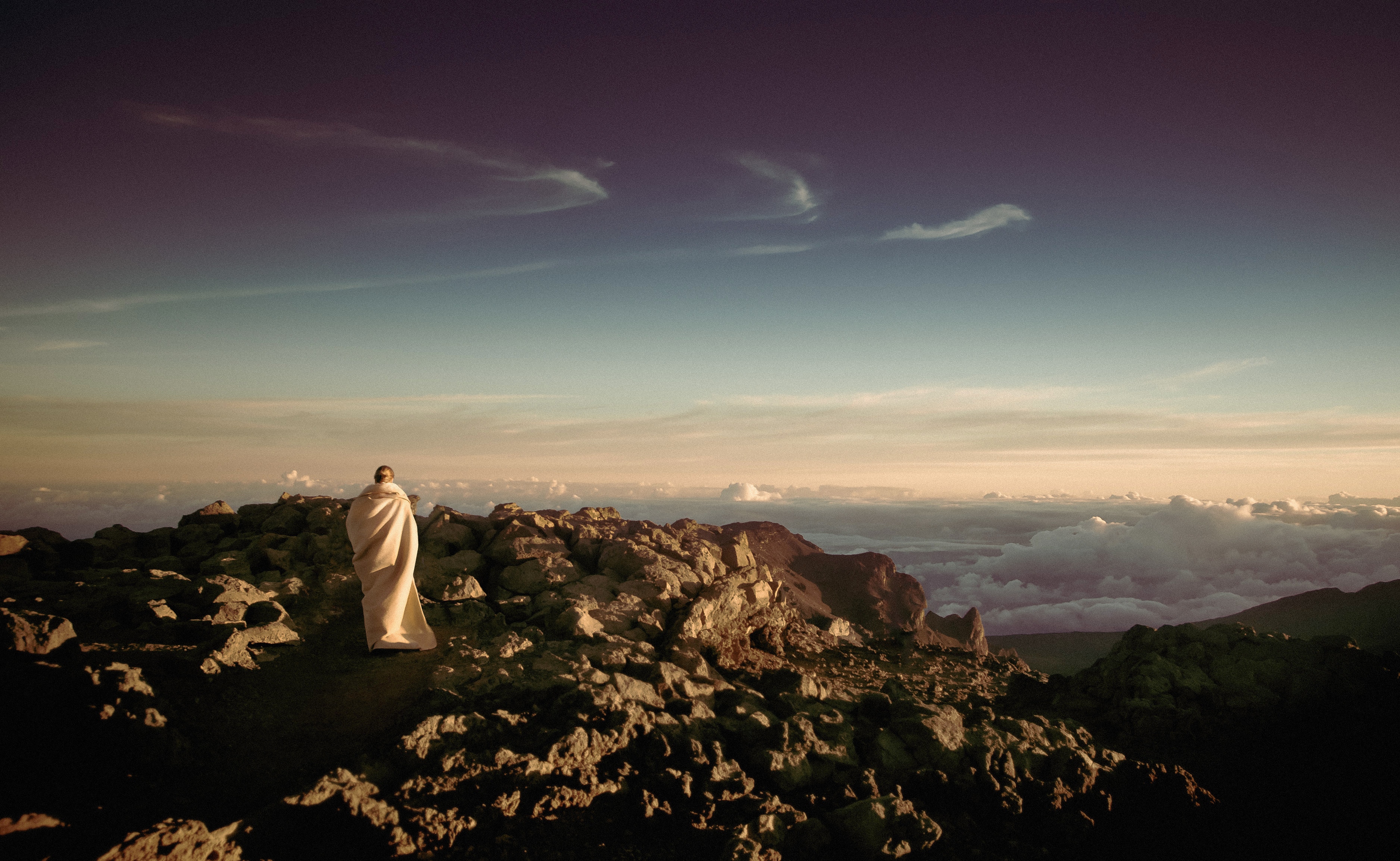 Person wrapped in a blanket on a rocky mountain peak HD Wallpaper