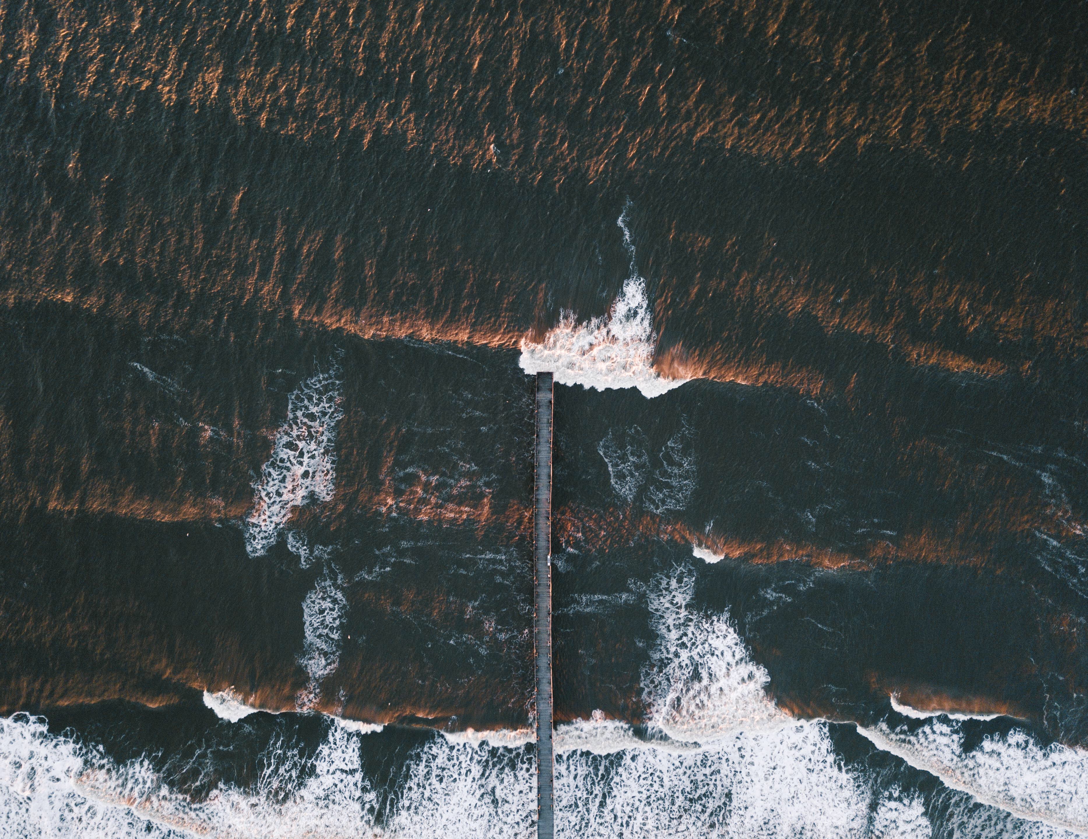 Aerial view of a pier extending into the ocean with waves HD Wallpaper