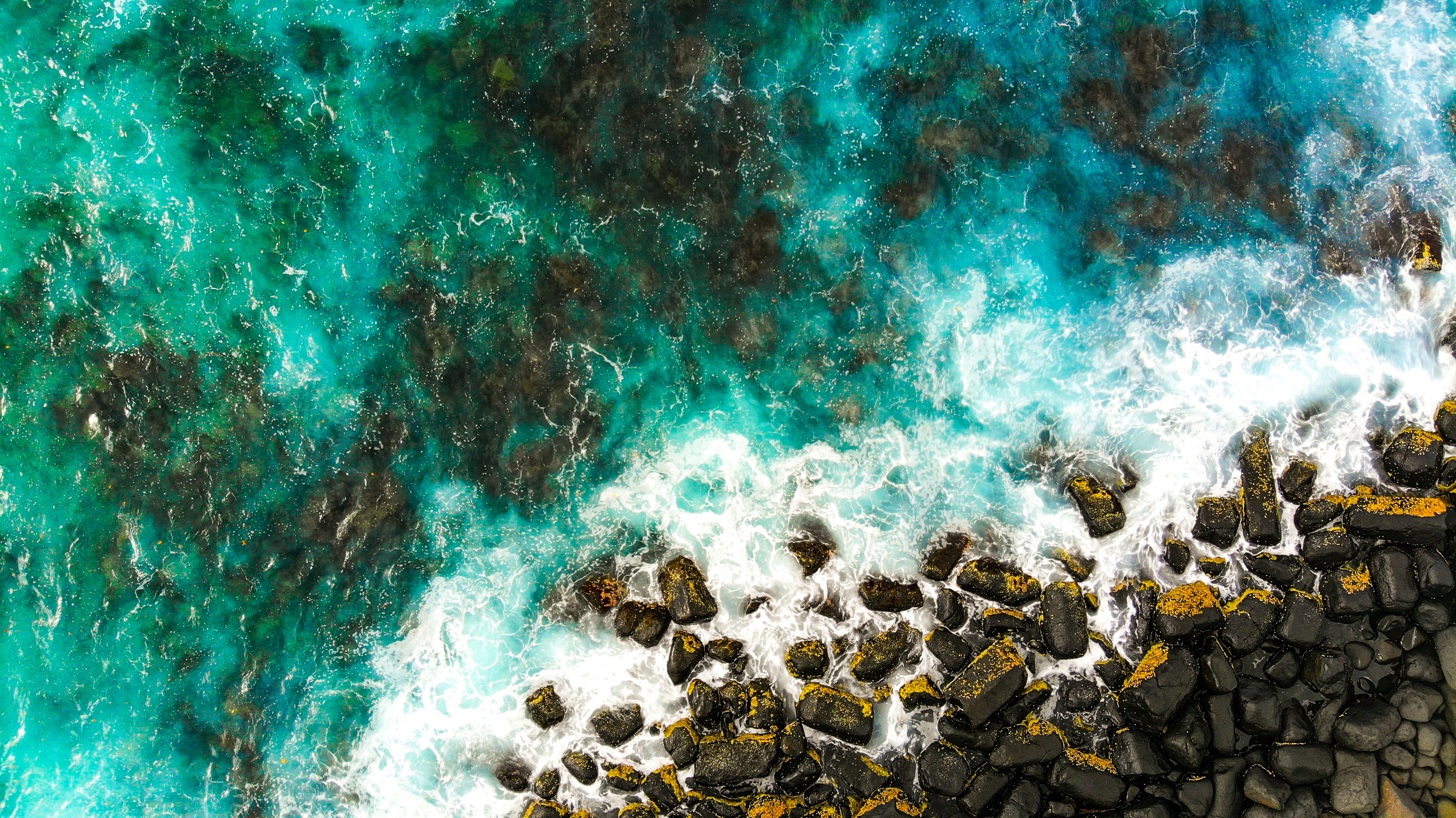 Aerial view of turquoise ocean waves crashing on a rocky coastline HD Wallpaper