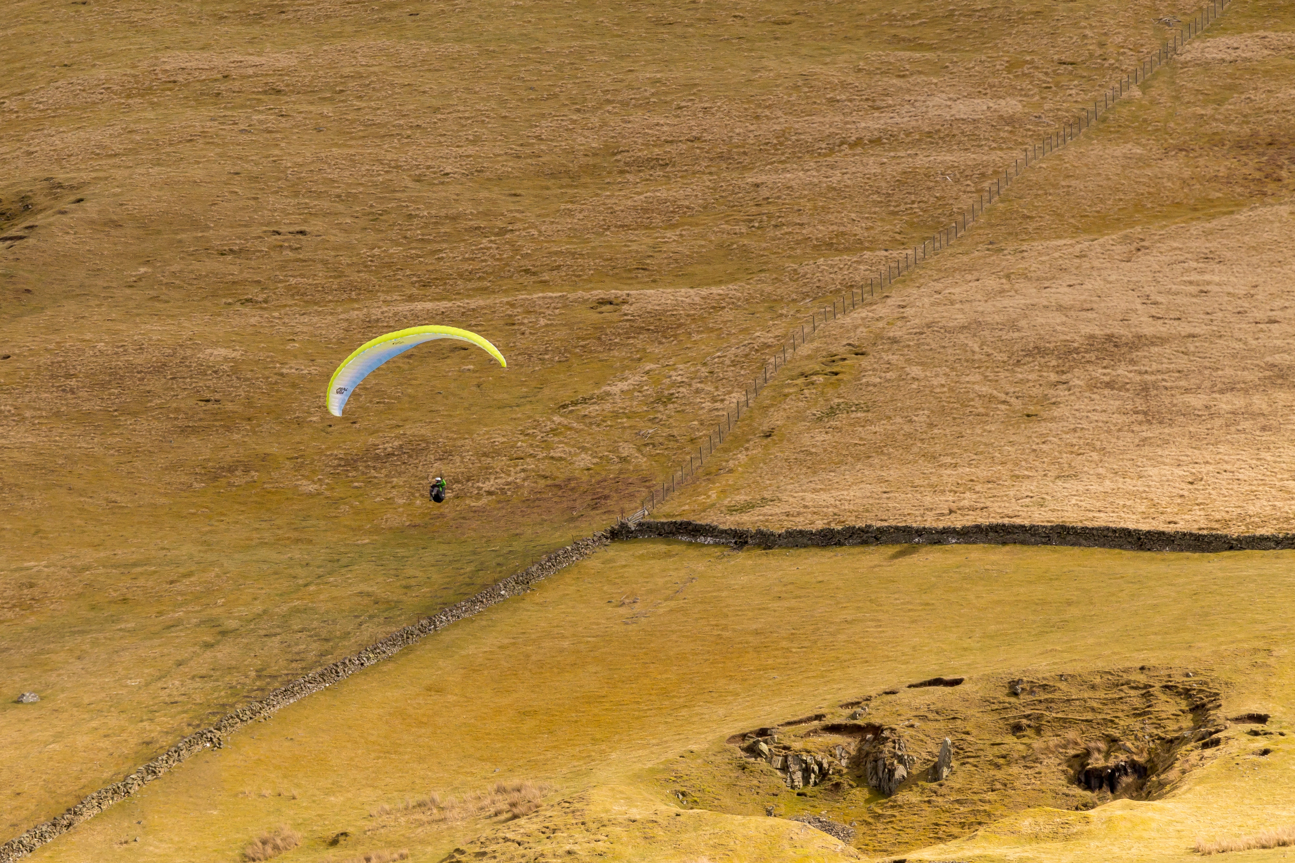 Paraglider flying over a dry, grassy field with stone walls HD Wallpaper