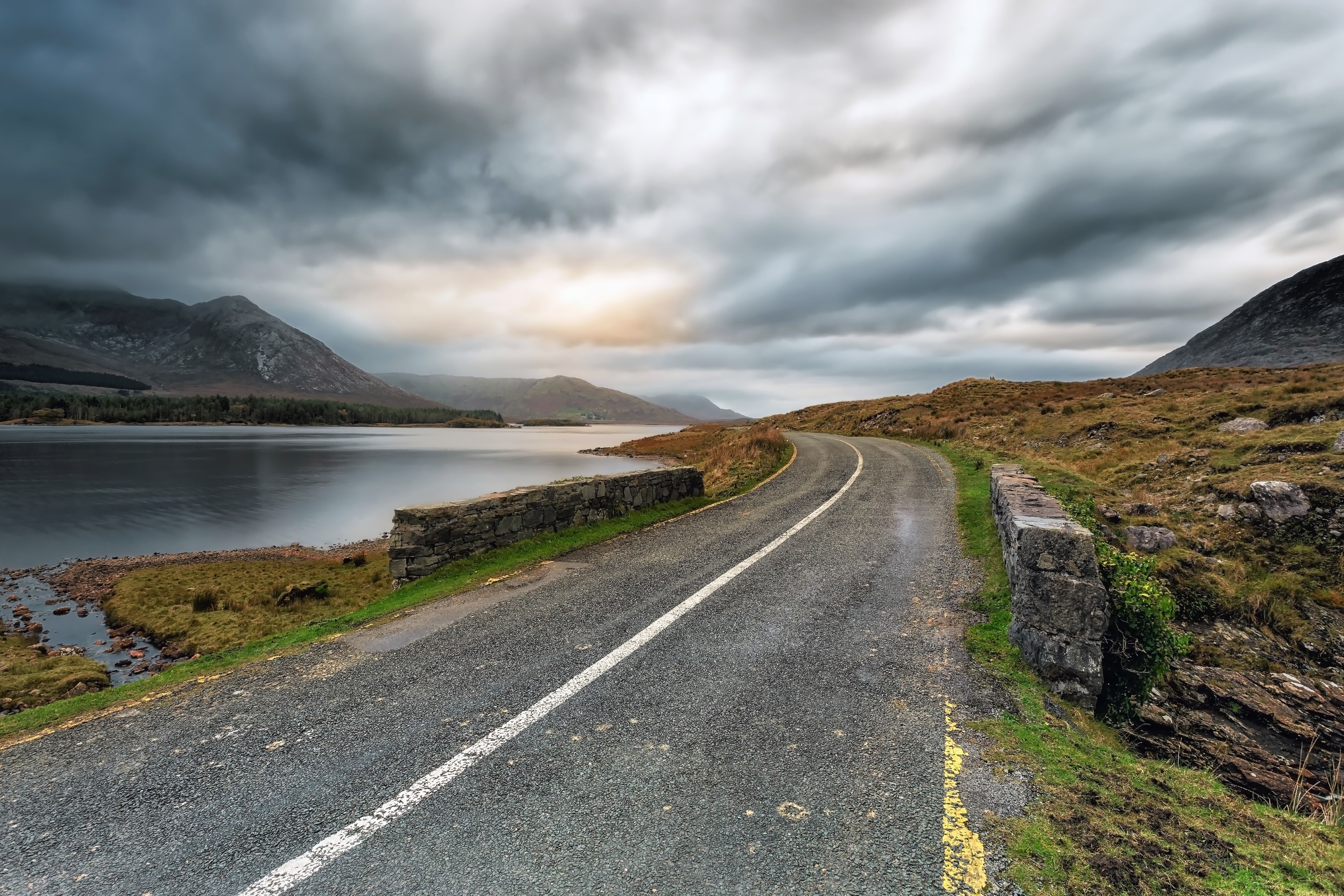 Road by a lake with mountains under cloudy sky 4K HD Wallpaper