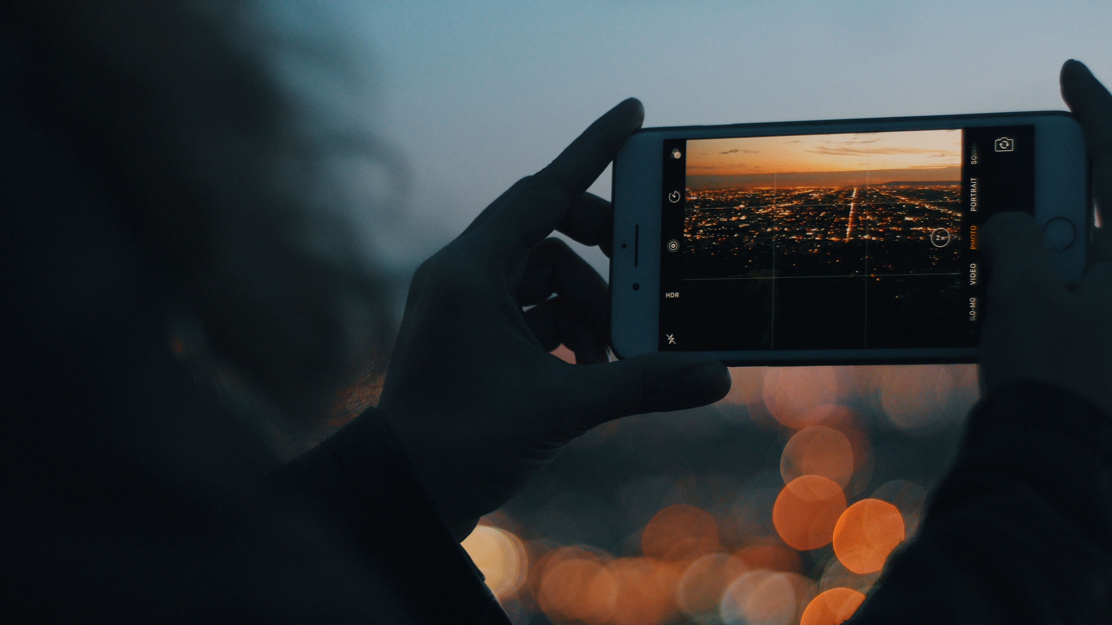 Person holding iPhone capturing city lights at dusk HD Wallpaper