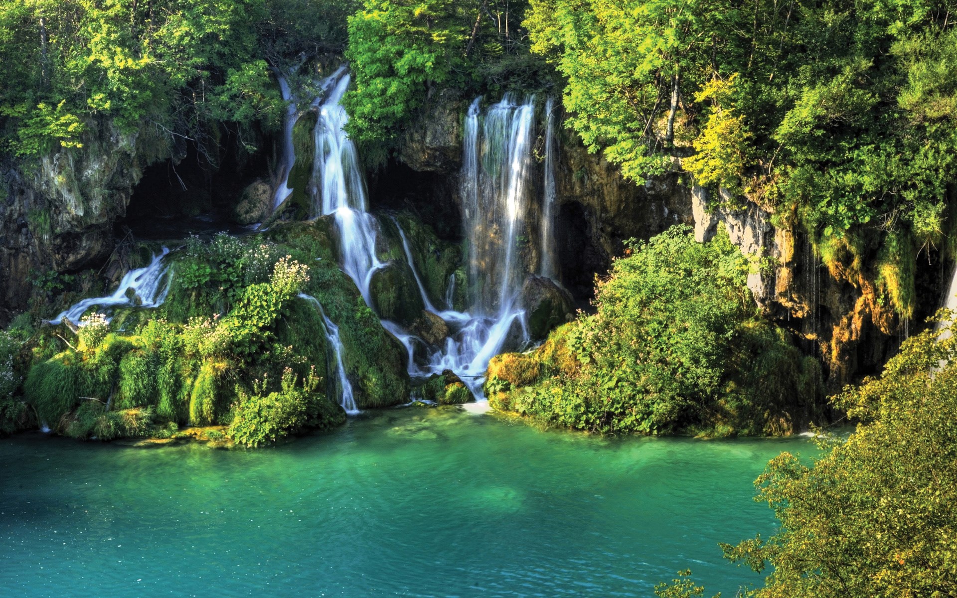 Waterfalls cascading into a turquoise lake surrounded by greenery HD Wallpaper