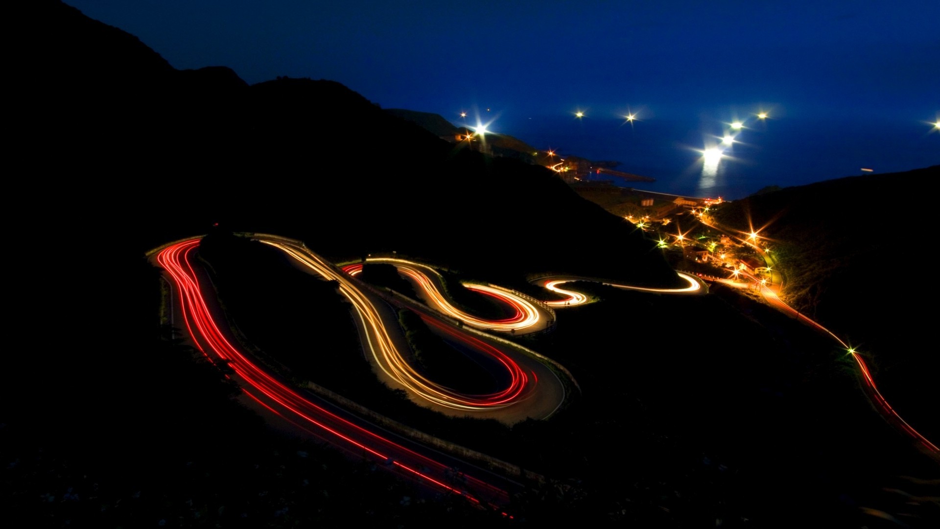 Night road with light trails and coastal town HD Wallpaper