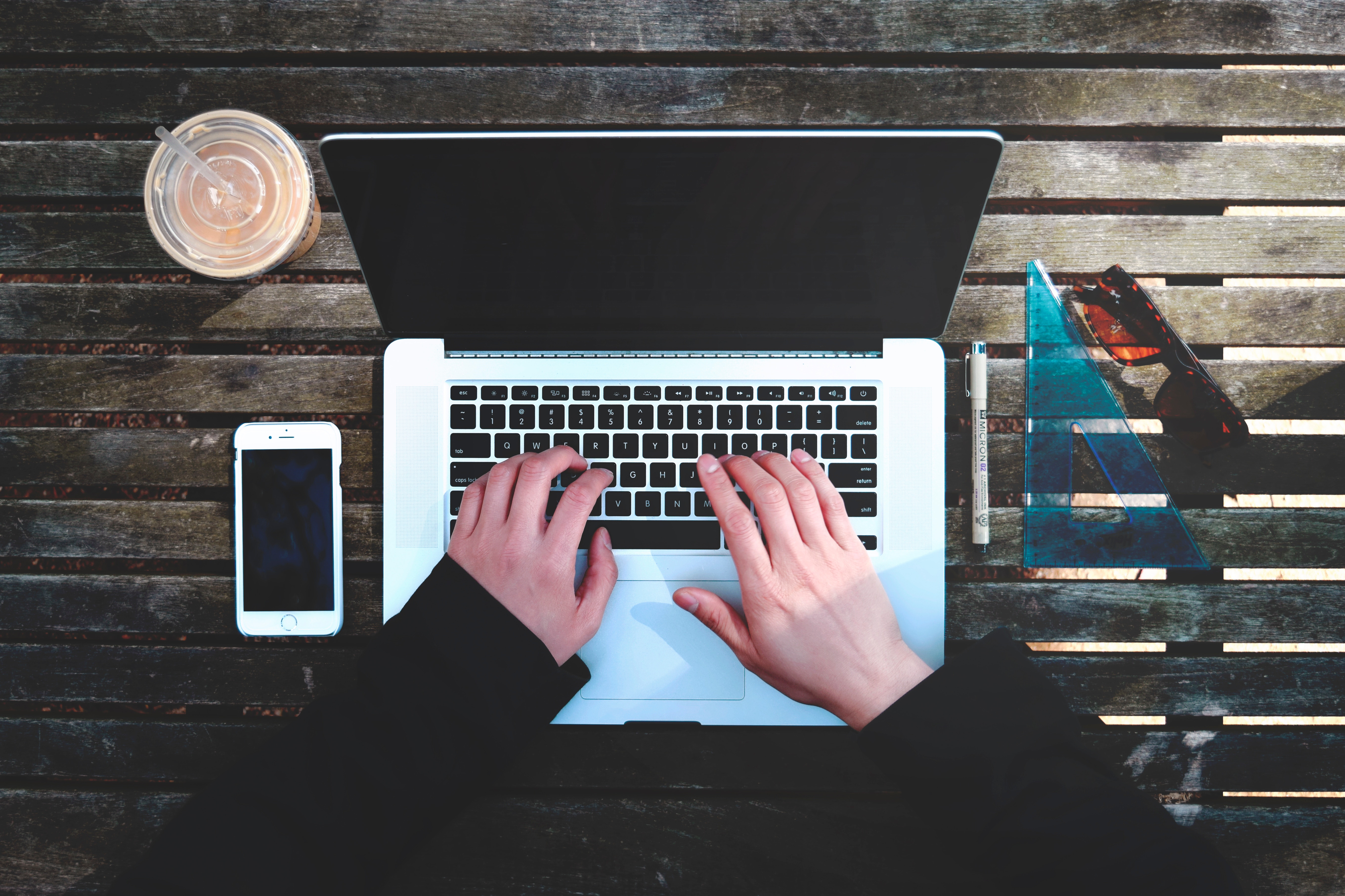 Hands typing on a MacBook with iPhone and iced coffee HD Wallpaper