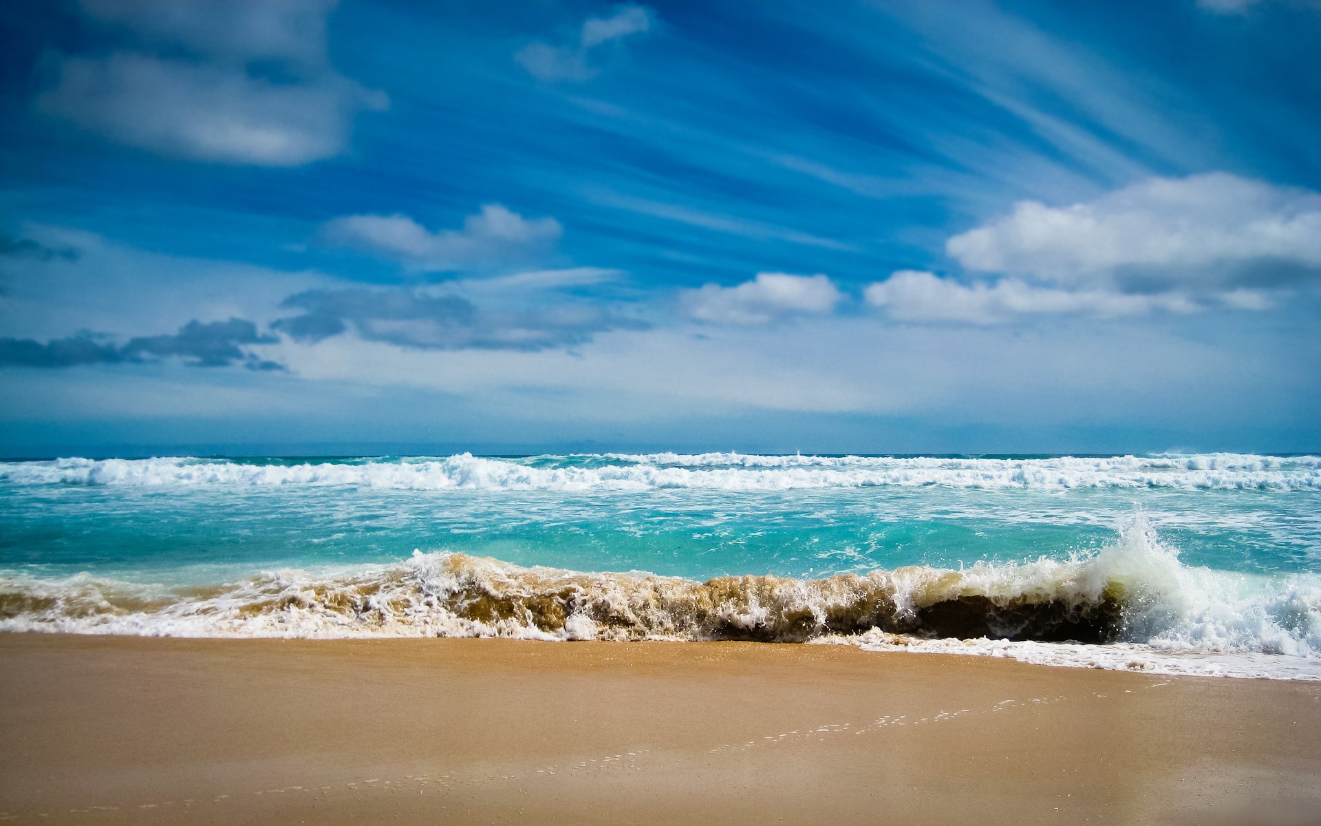 Ocean waves crashing on a sandy beach under a cloudy blue sky HD Wallpaper