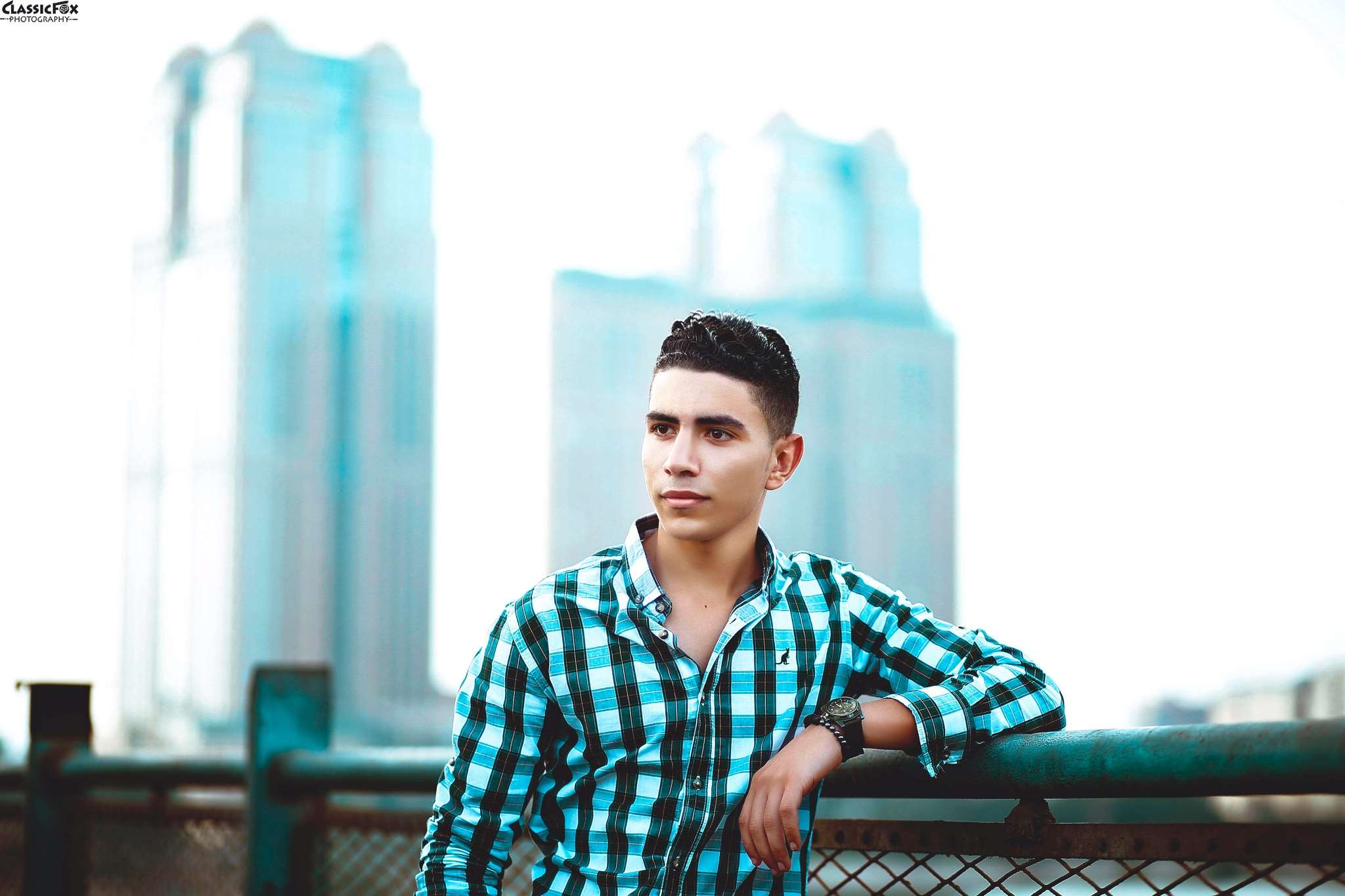 Young man in plaid shirt leaning on fence with buildings HD Wallpaper