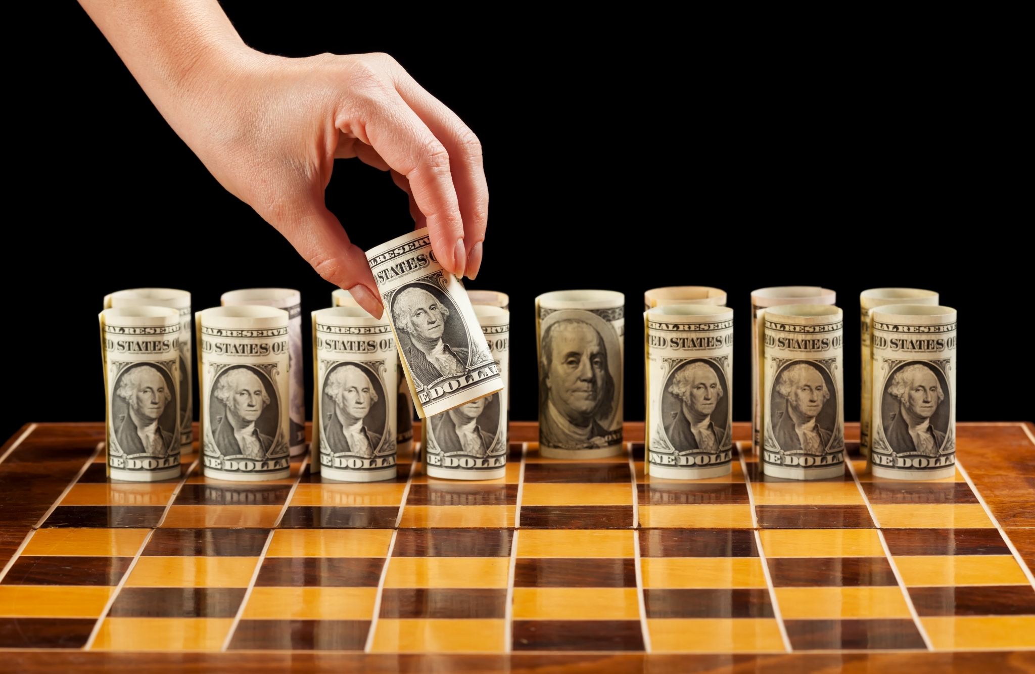 Hand placing rolled dollar bills on a chessboard HD Wallpaper