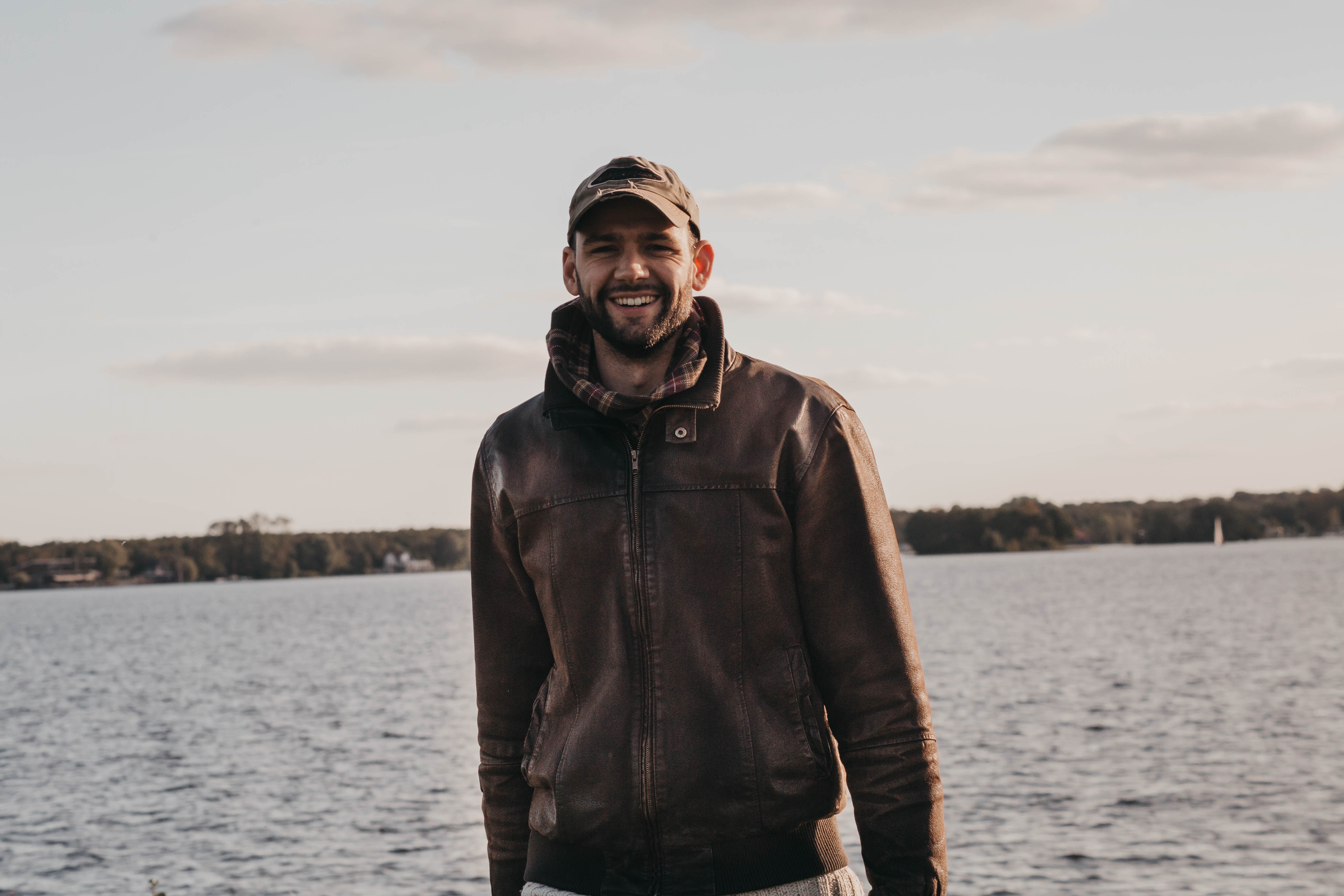 Man in leather jacket and cap smiling by the water HD Wallpaper