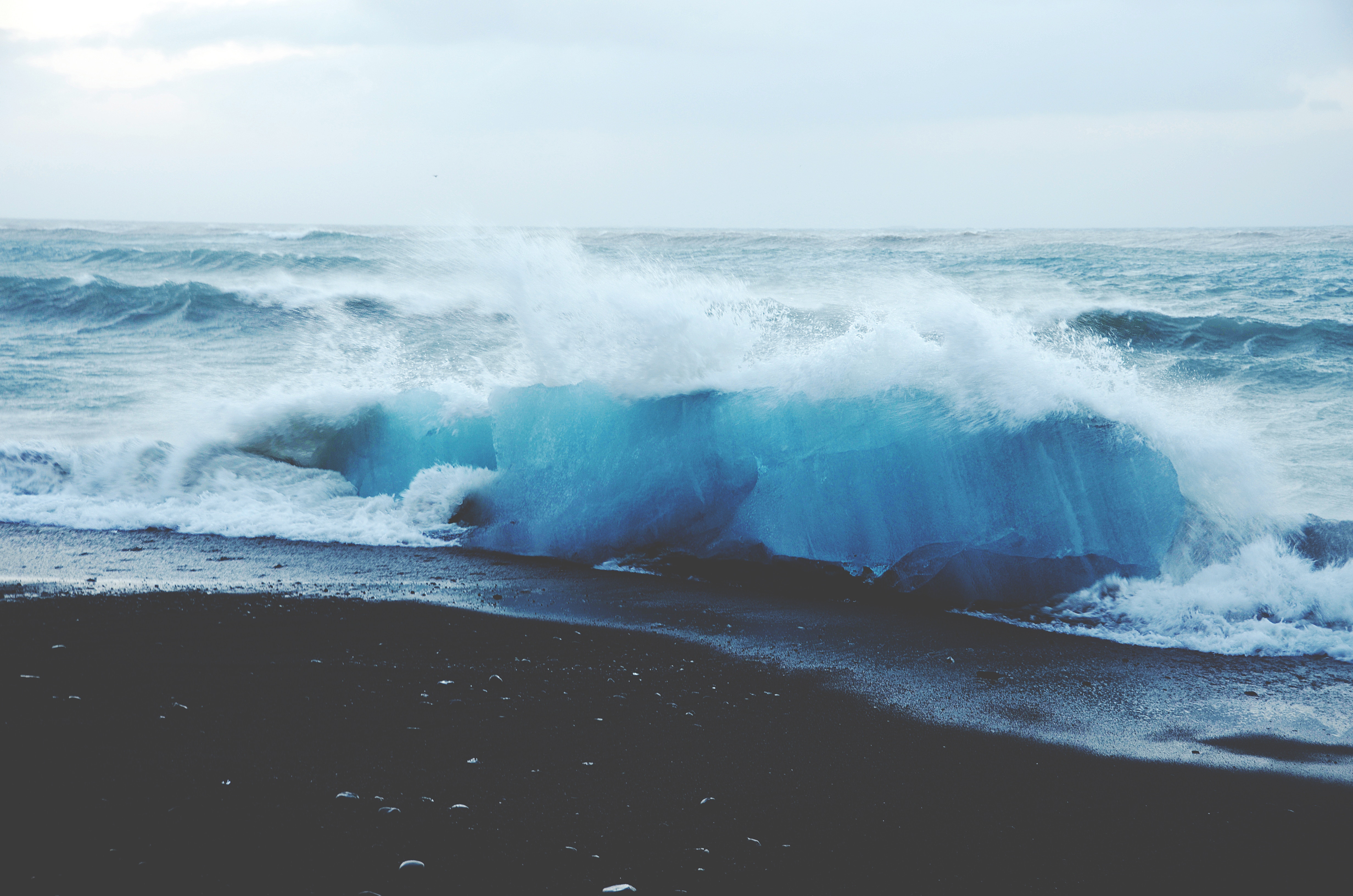 Blue iceberg on a black sand beach with crashing waves HD Wallpaper