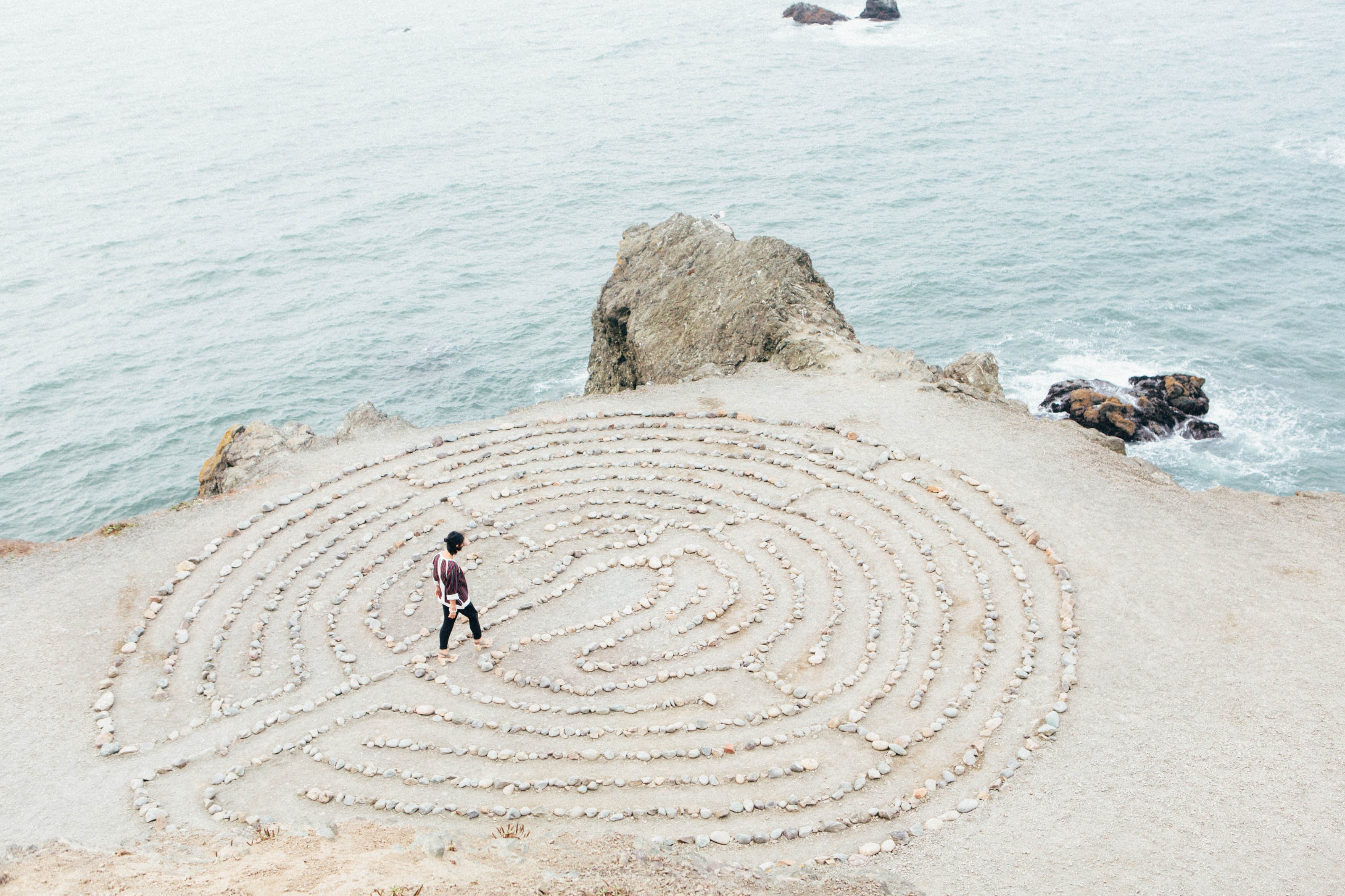 Person walking through a stone maze on a cliff overlooking the ocean HD Wallpaper