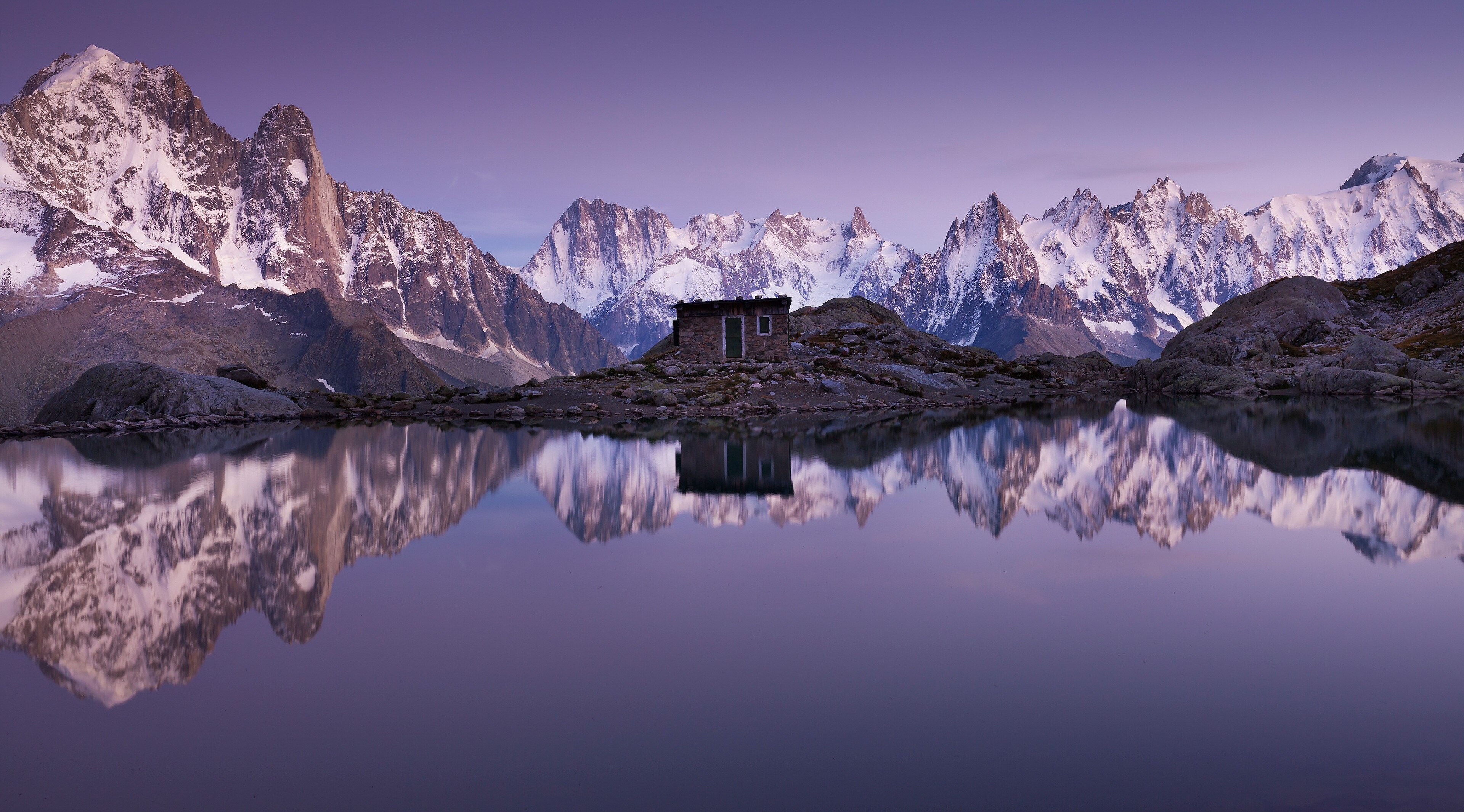 Snowy mountains reflected in a lake with a small cabin HD Wallpaper