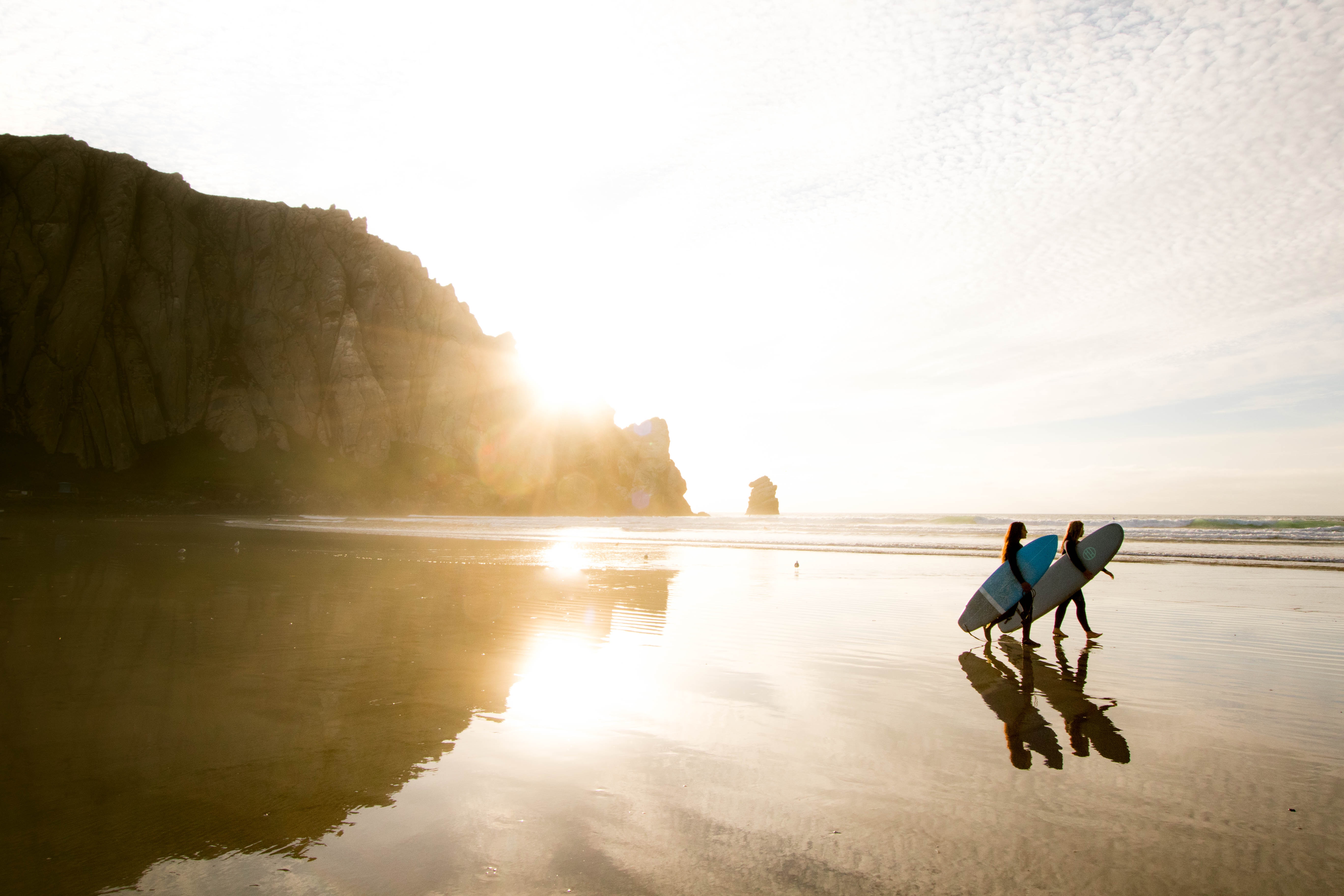 Two surfers walk along a beach at sunset with a cliff in the background. HD Wallpaper