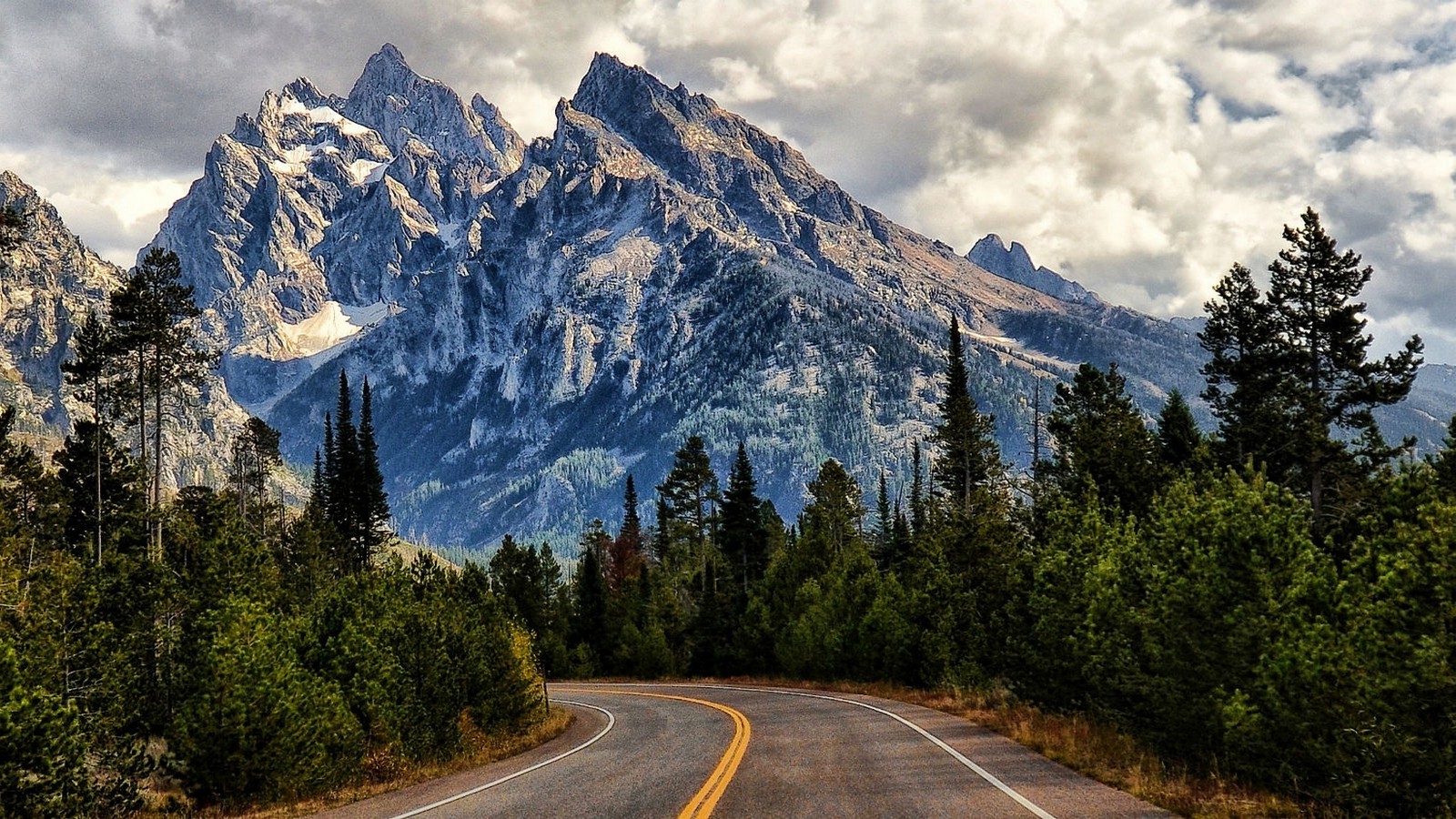 Mountain road through a forest with snow-capped peaks HD Wallpaper