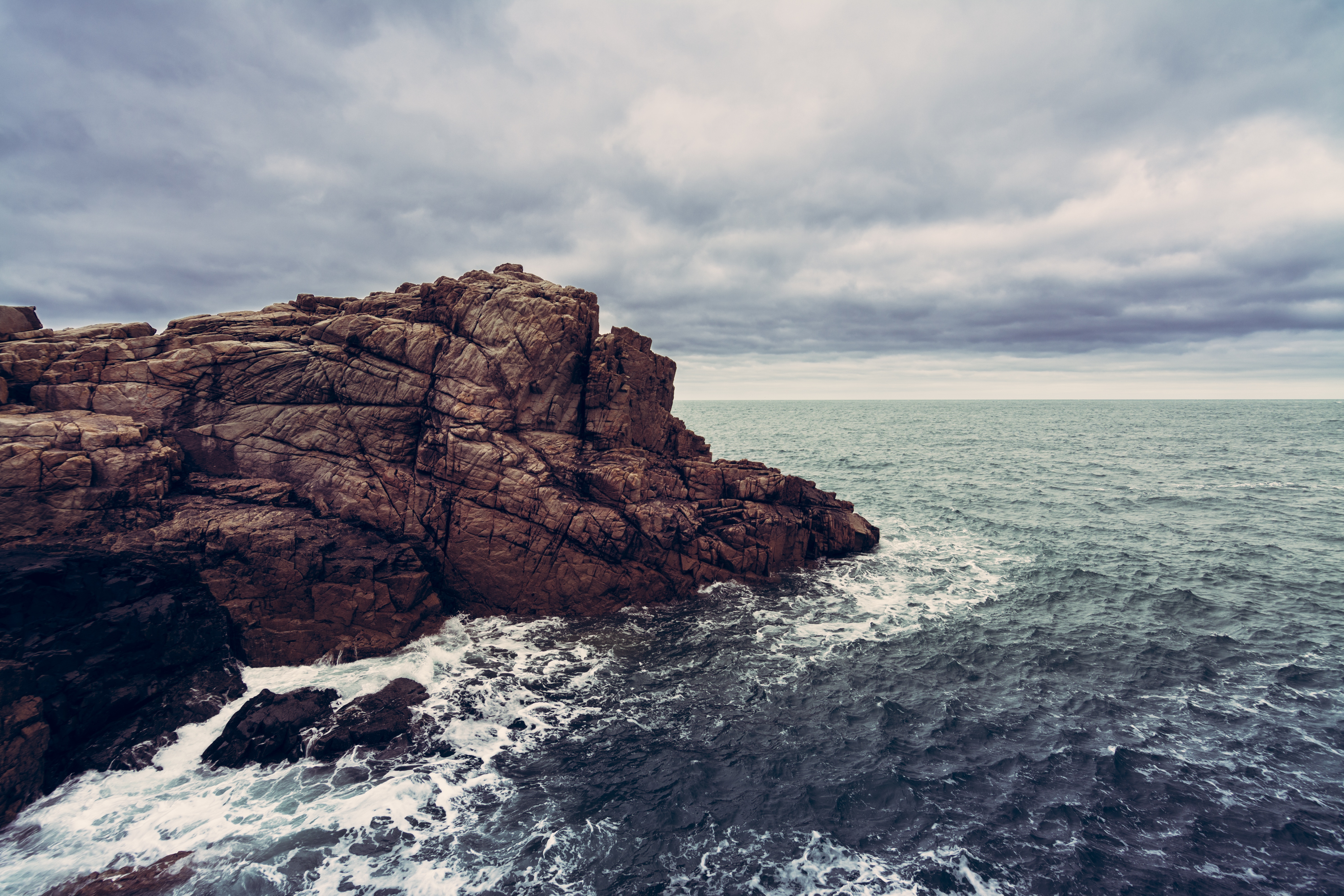 Brown rocky cliff meets the ocean under a cloudy sky HD Wallpaper