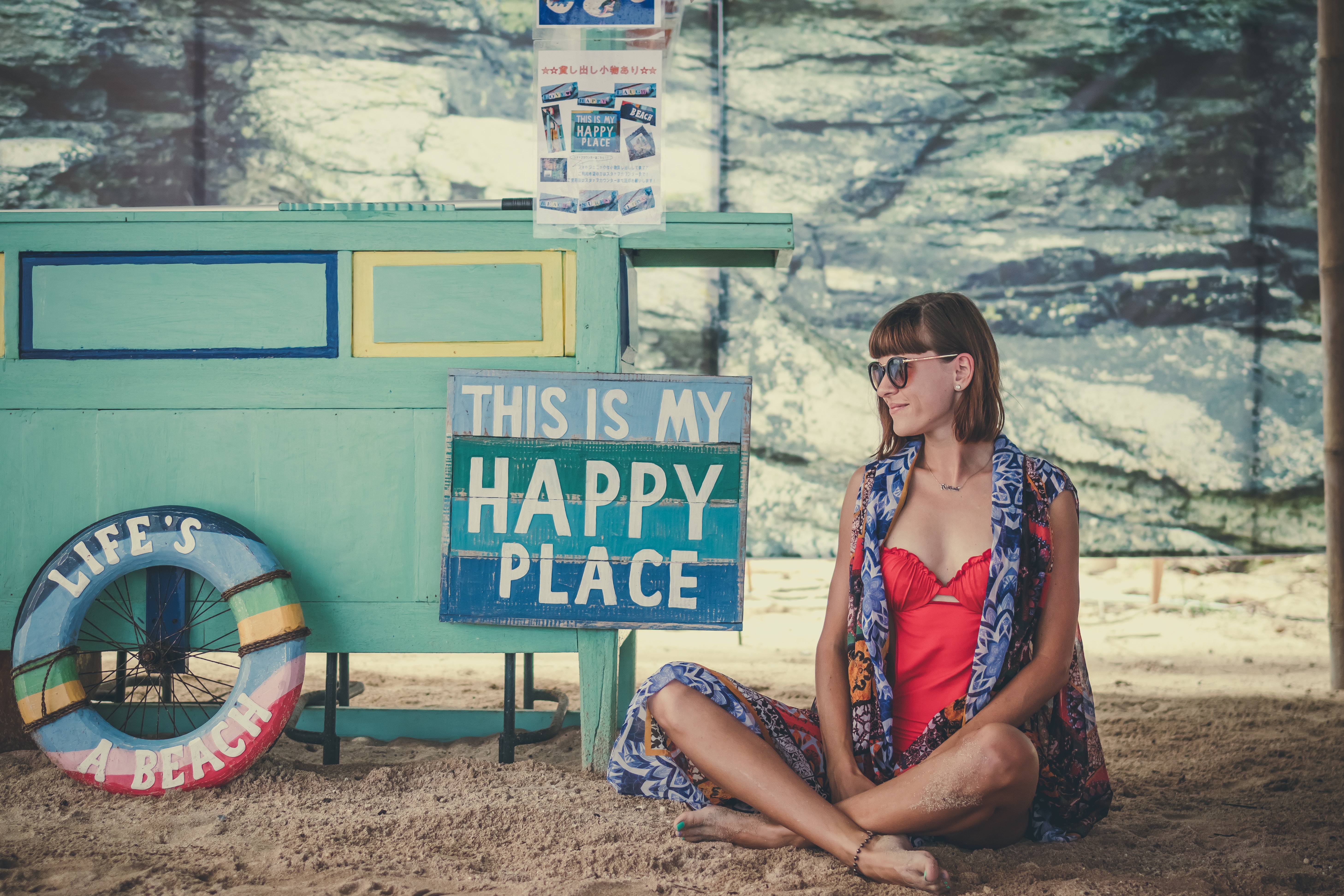 Woman in red swimsuit sits on sand near a cart and sign HD Wallpaper