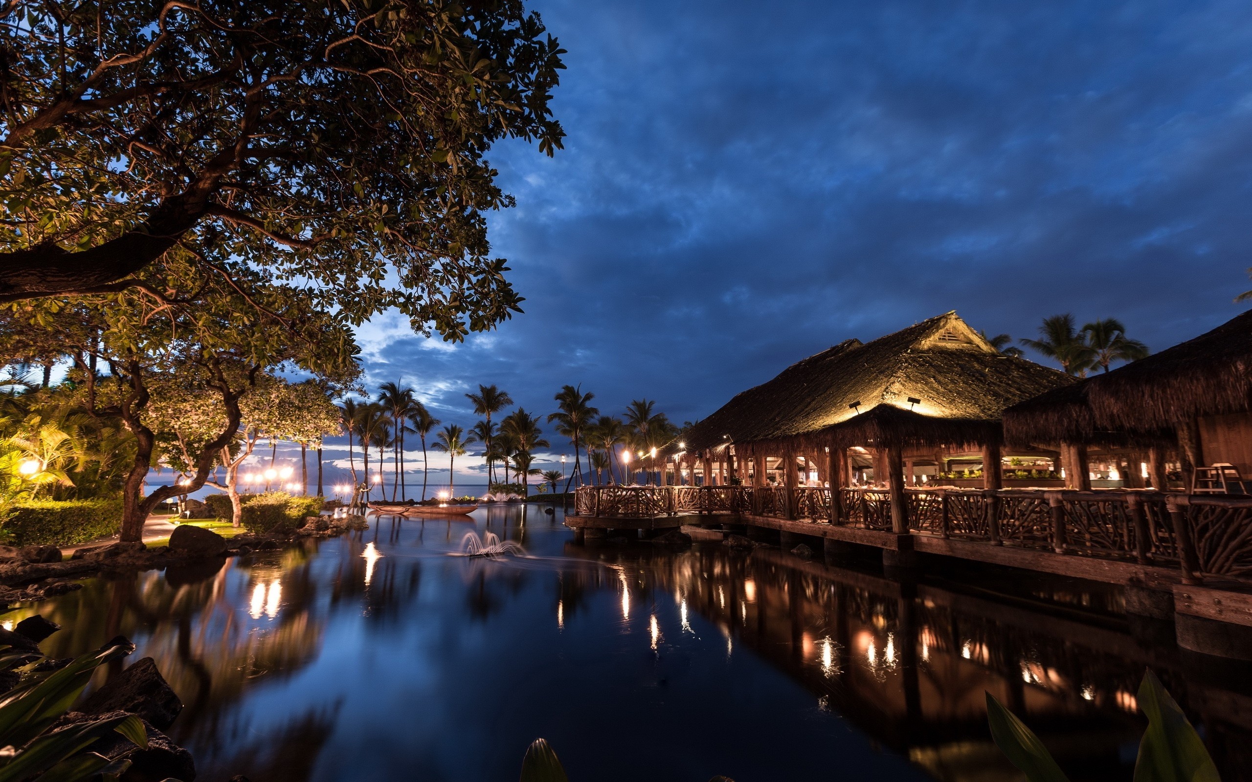 Tropical resort at night with thatched roof buildings and a pond HD Wallpaper