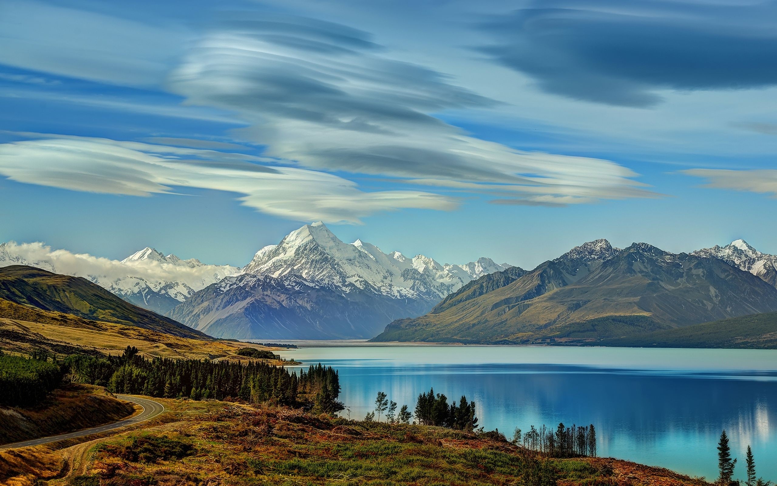 Snow-capped mountains reflected in a blue lake with a winding road HD Wallpaper