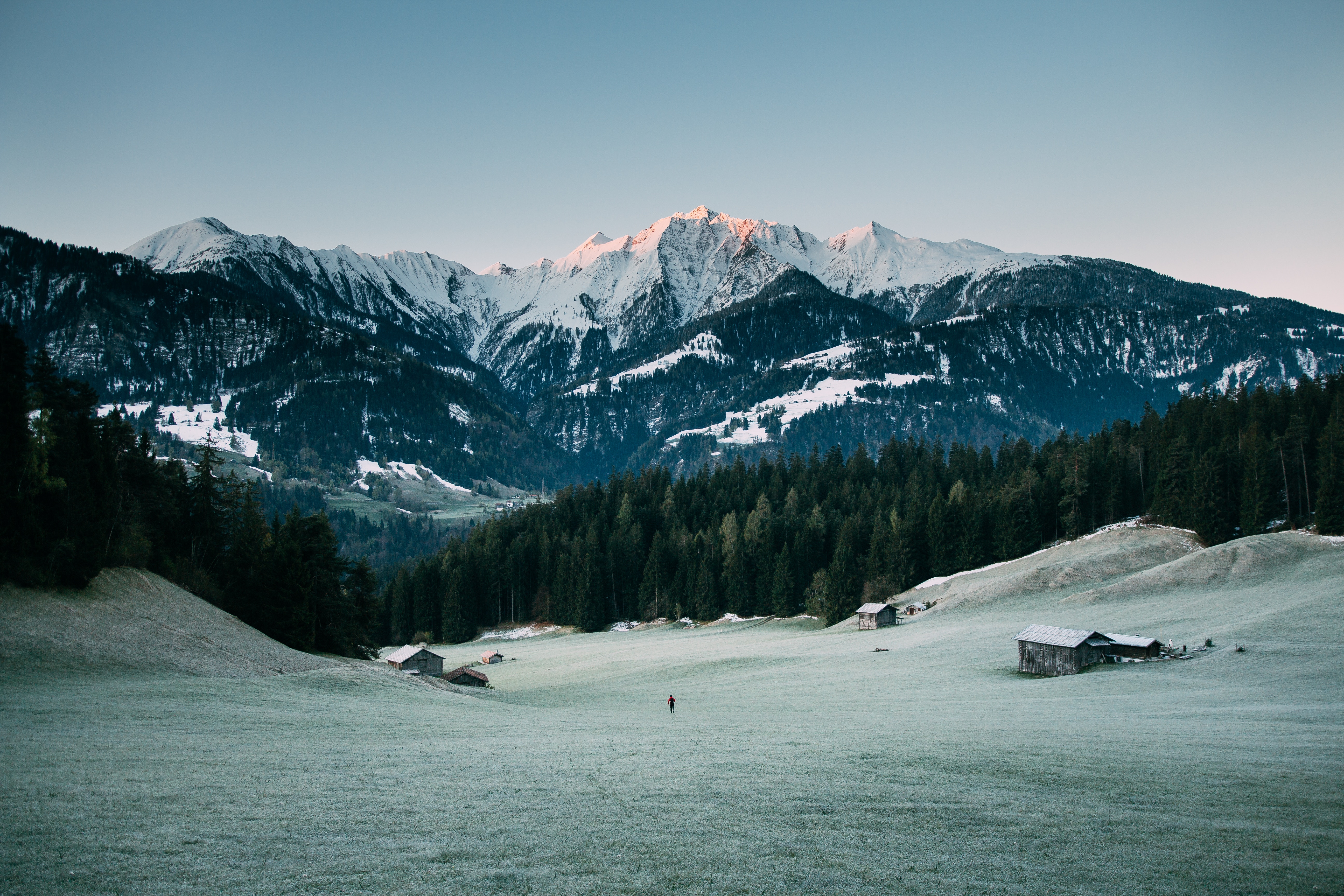 Lone person walks through a frosty field towards snow-capped mountains at sunrise HD Wallpaper