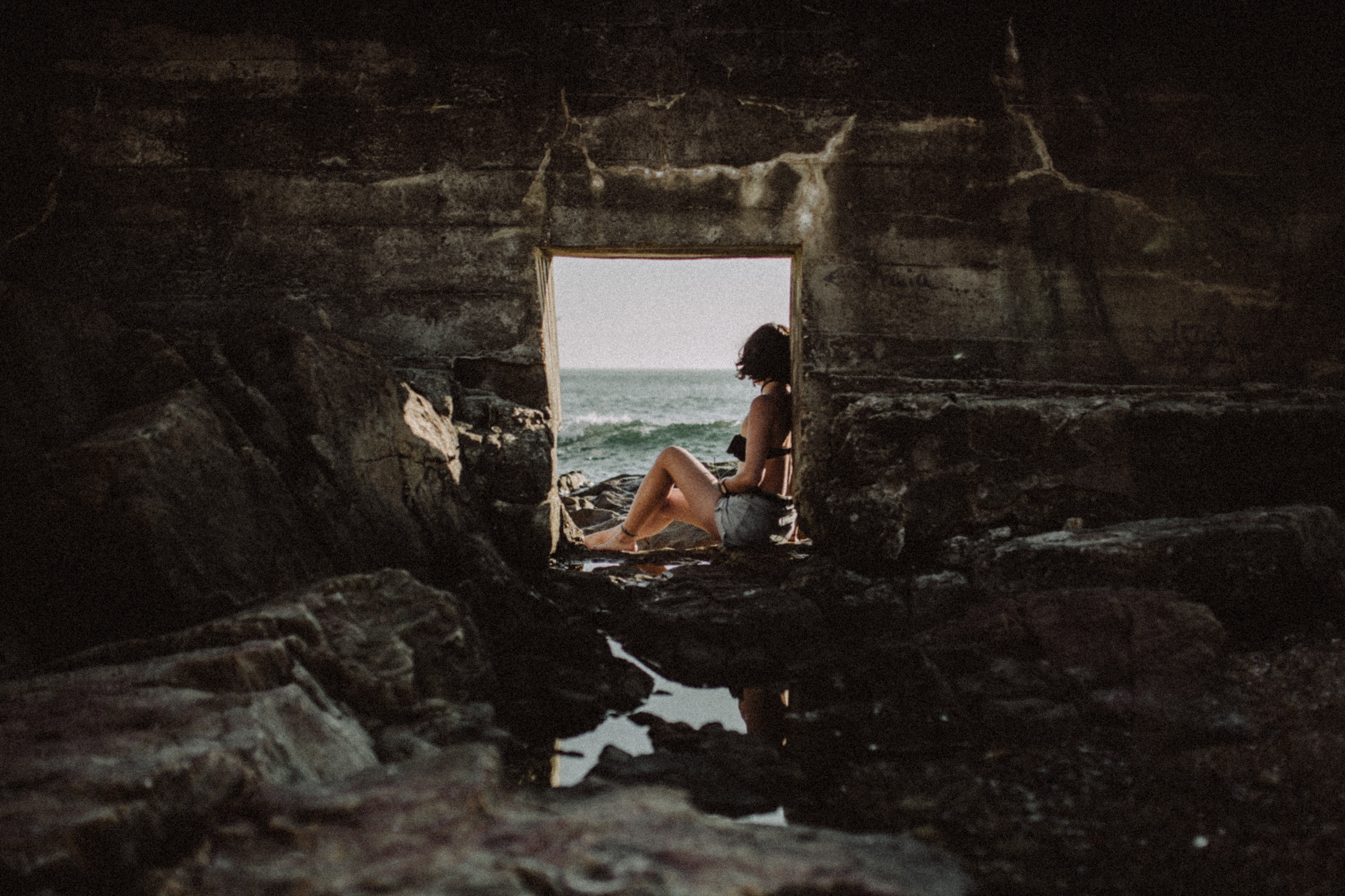 Woman sitting in a stone cave looking at the ocean HD Wallpaper