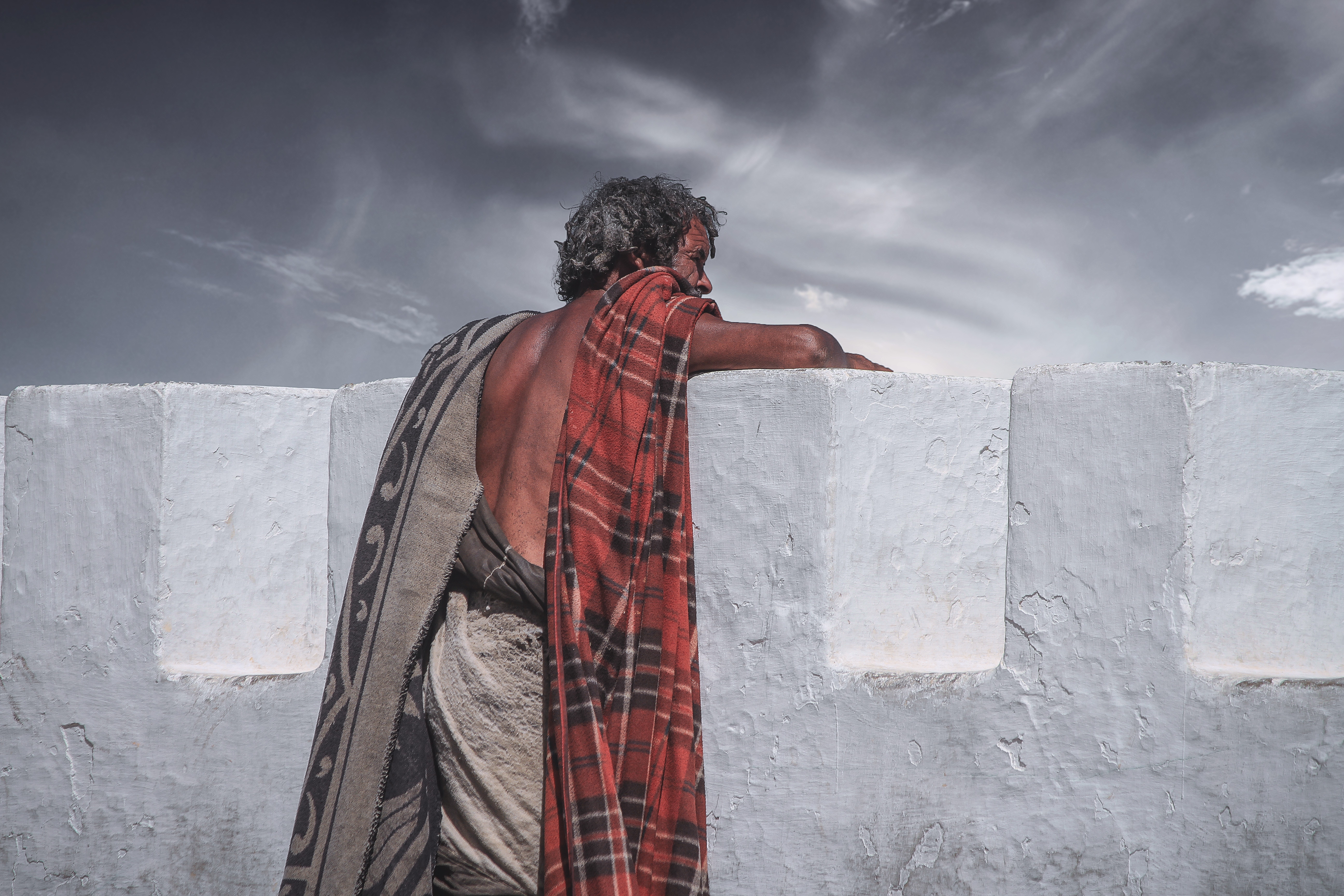 Man in blankets leans on a white wall under a cloudy sky HD Wallpaper