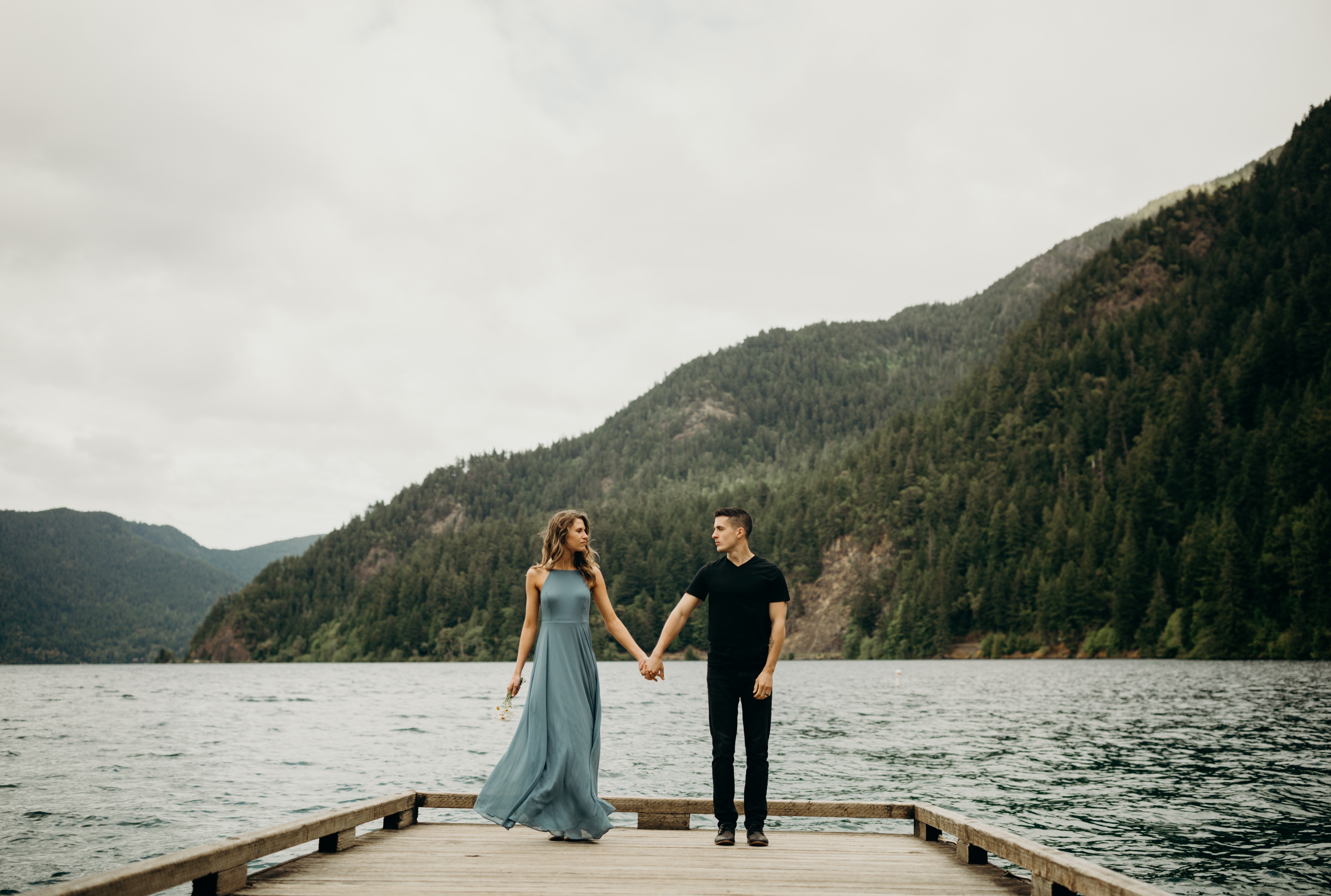 Couple holding hands on a wooden pier with mountains HD Wallpaper