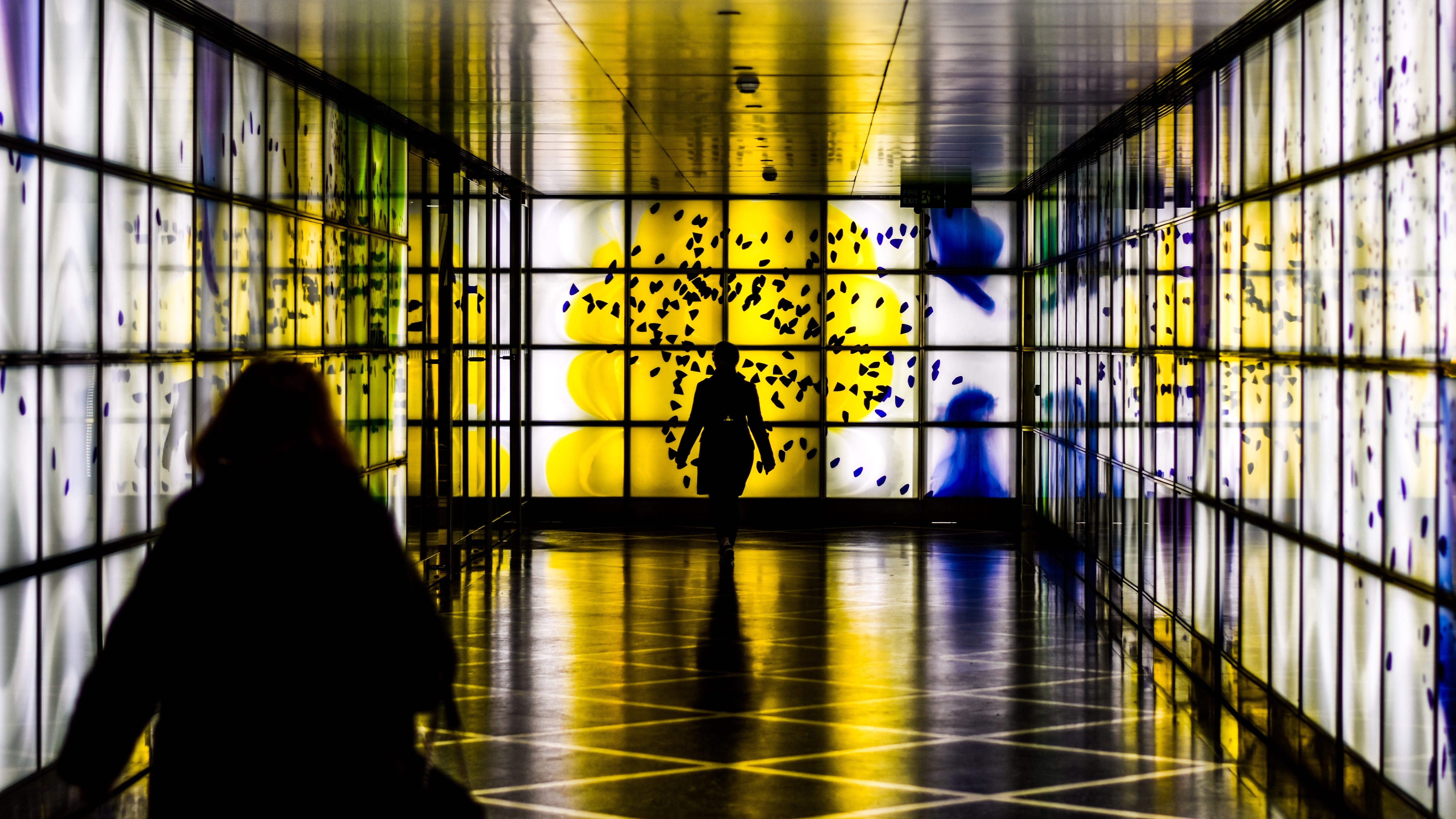 People walking through a brightly lit modern corridor with glowing panels HD Wallpaper
