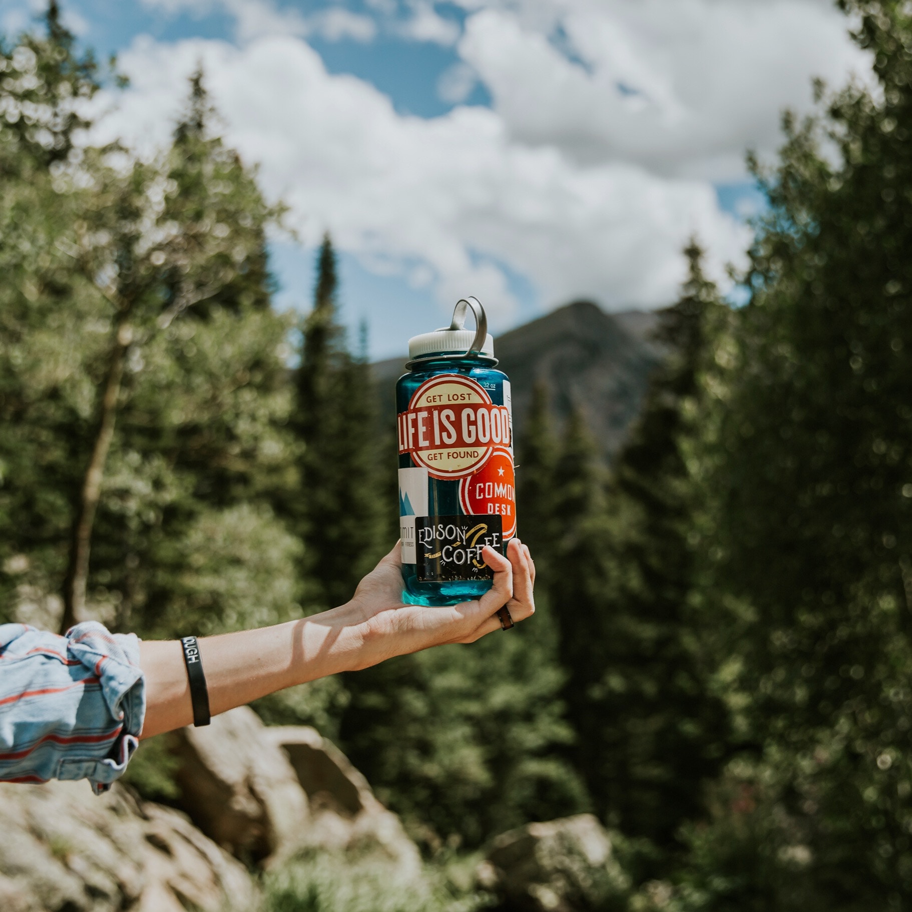 Person holding a "Life is Good" water bottle in a forest HD Wallpaper