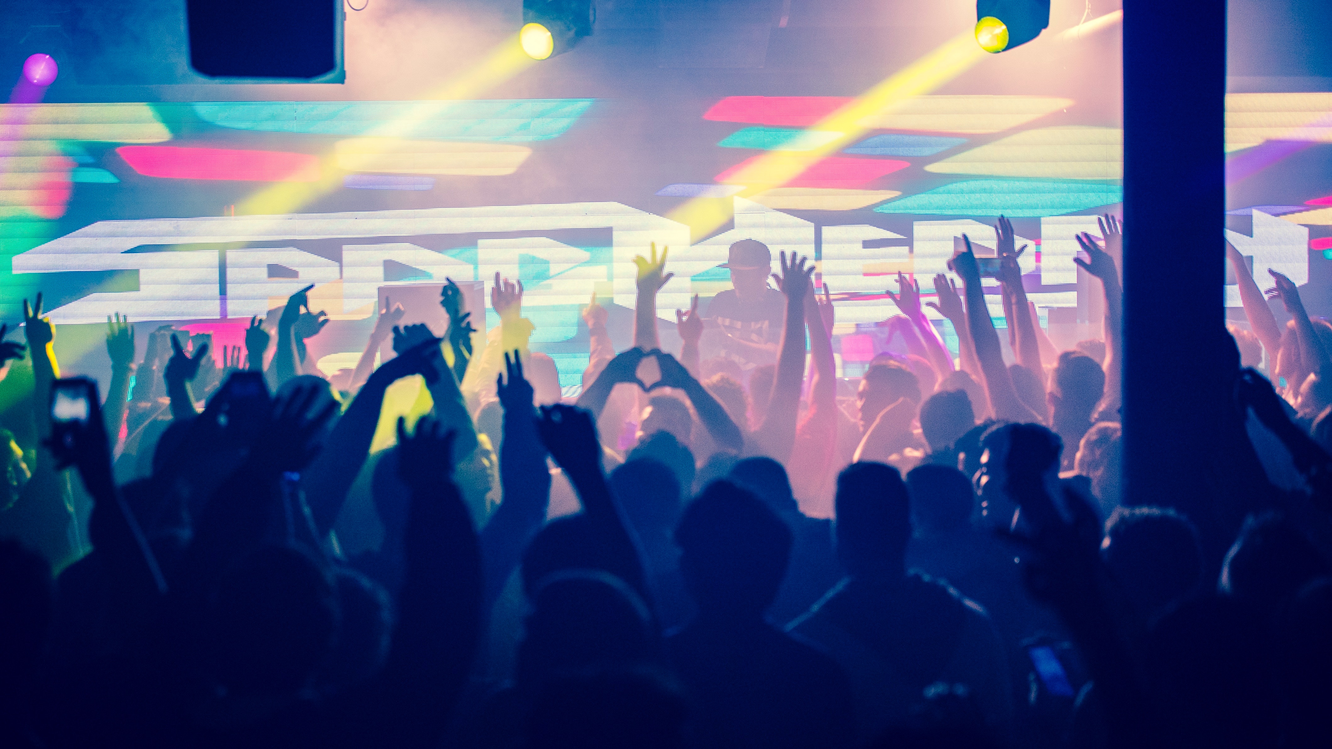 Crowd with arms raised at a club with colorful lights HD Wallpaper